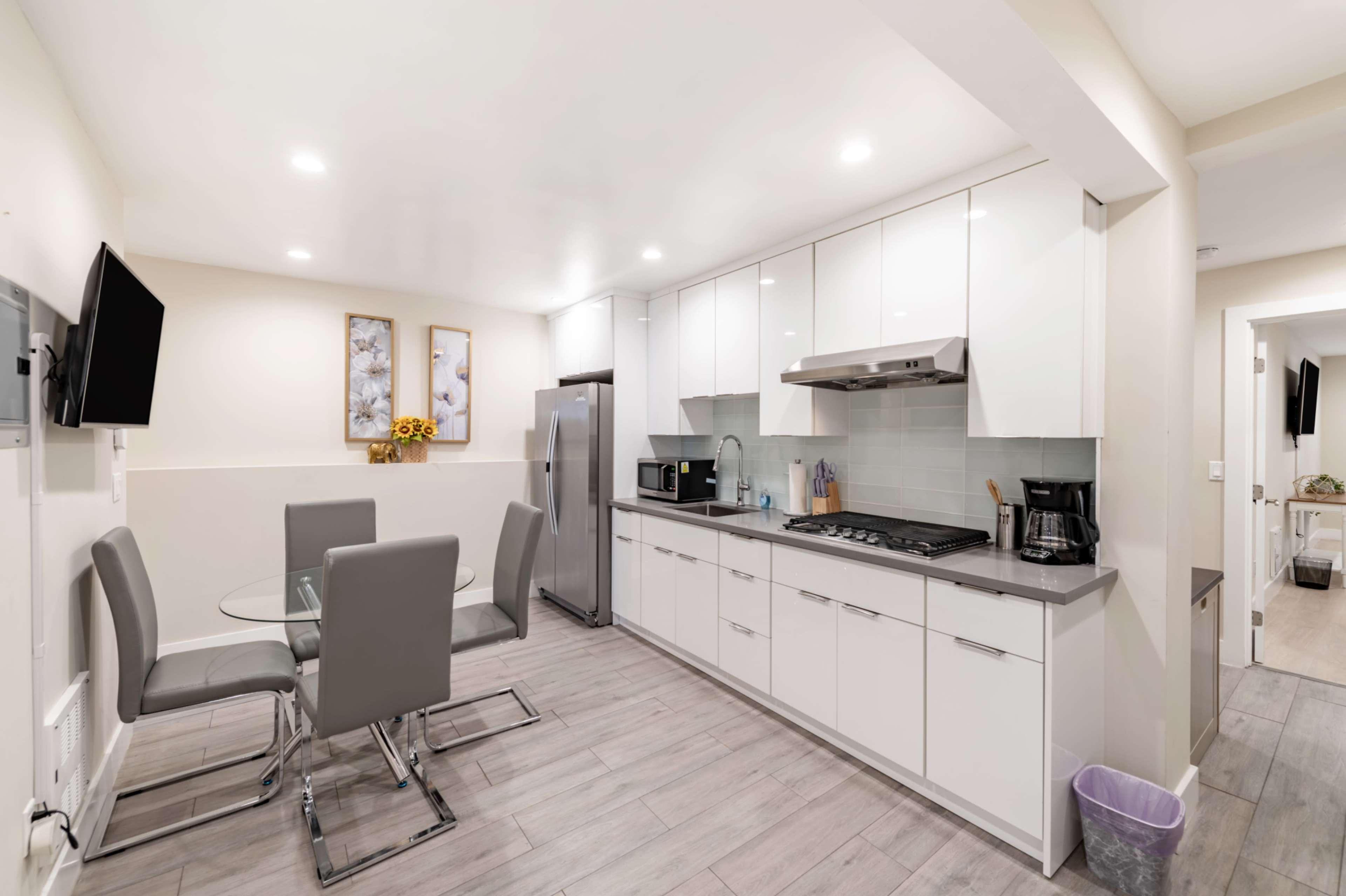 The image shows a modern kitchen with white cabinetry, stainless steel appliances, a glass dining table, and gray chairs.