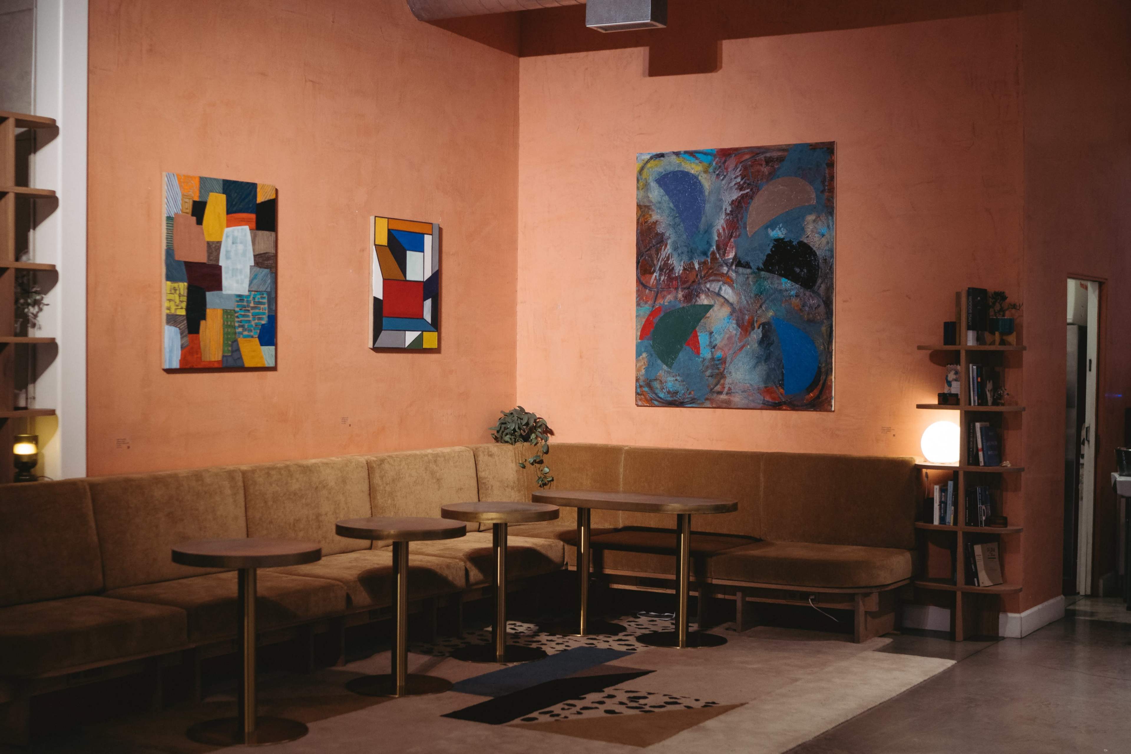 The image shows a cozy cafe setting with a brown upholstered seating area, two round tables, and colorful artwork hanging on the orange wall.