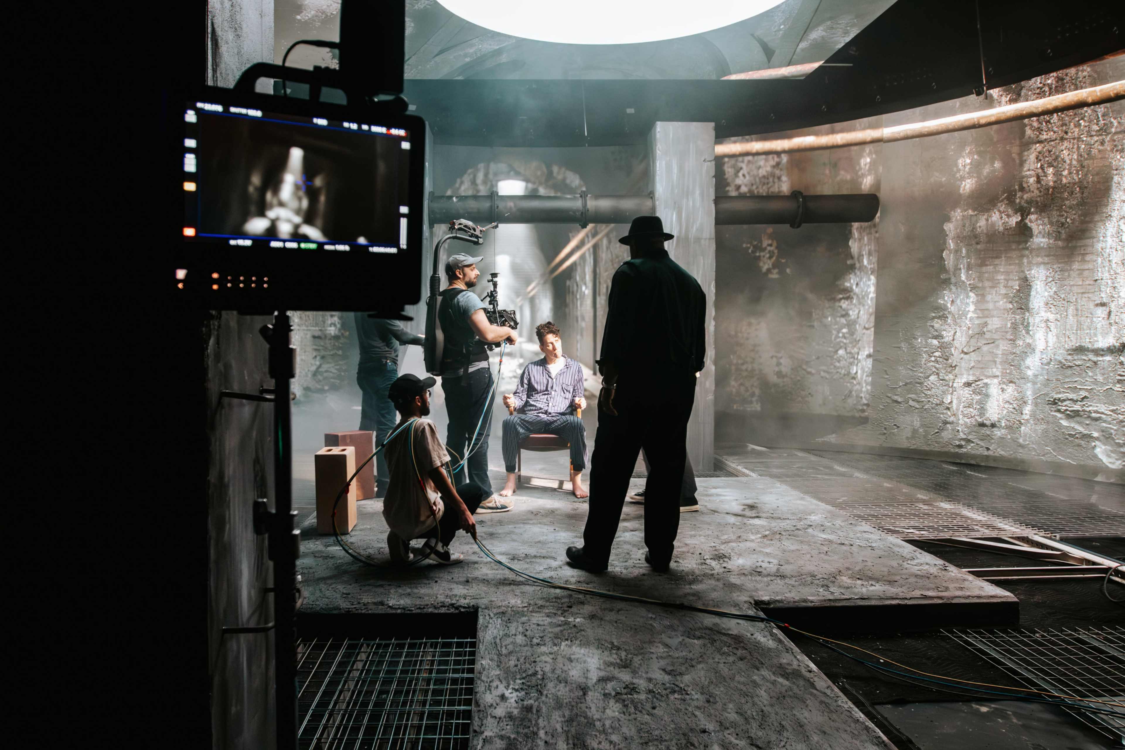 A film crew captures a scene in an industrial-style setting with smoke and dramatic lighting, featuring an actor seated in a chair.