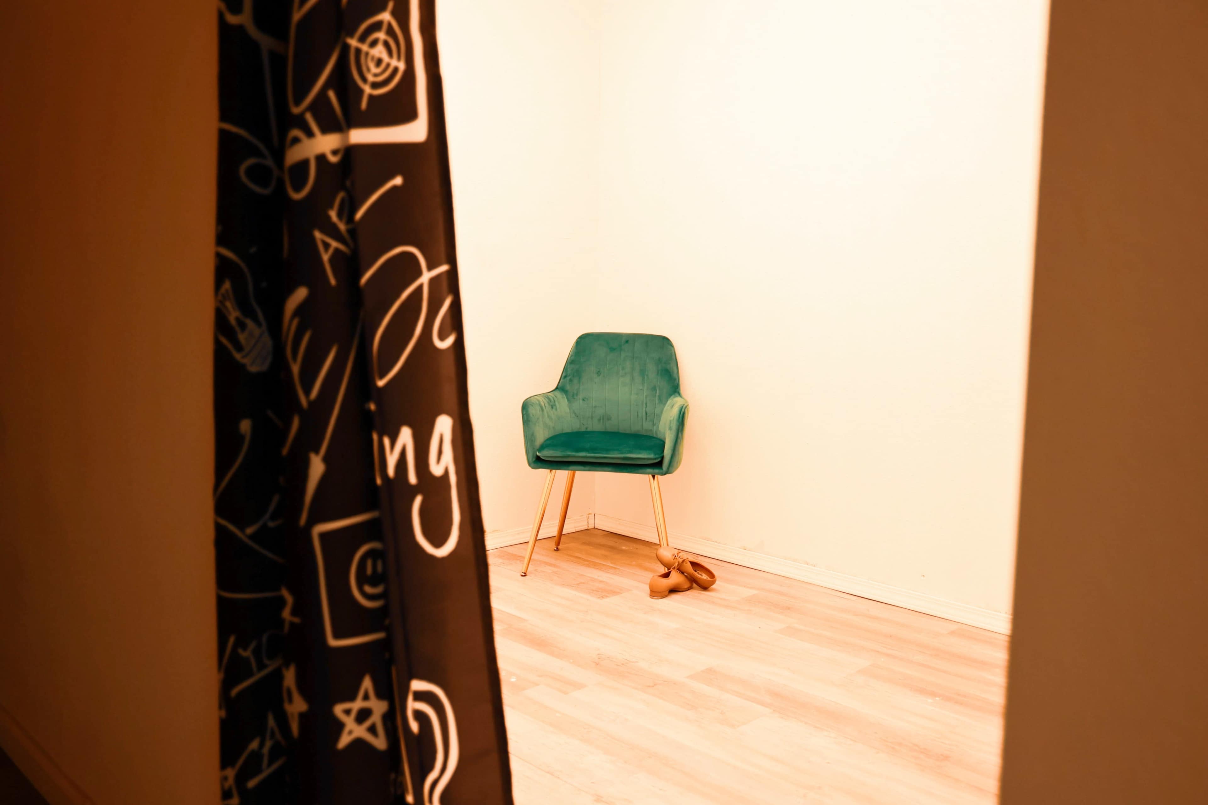 A green chair is positioned in a softly lit room, with a pair of shoes on the floor nearby and a patterned curtain partially drawn.