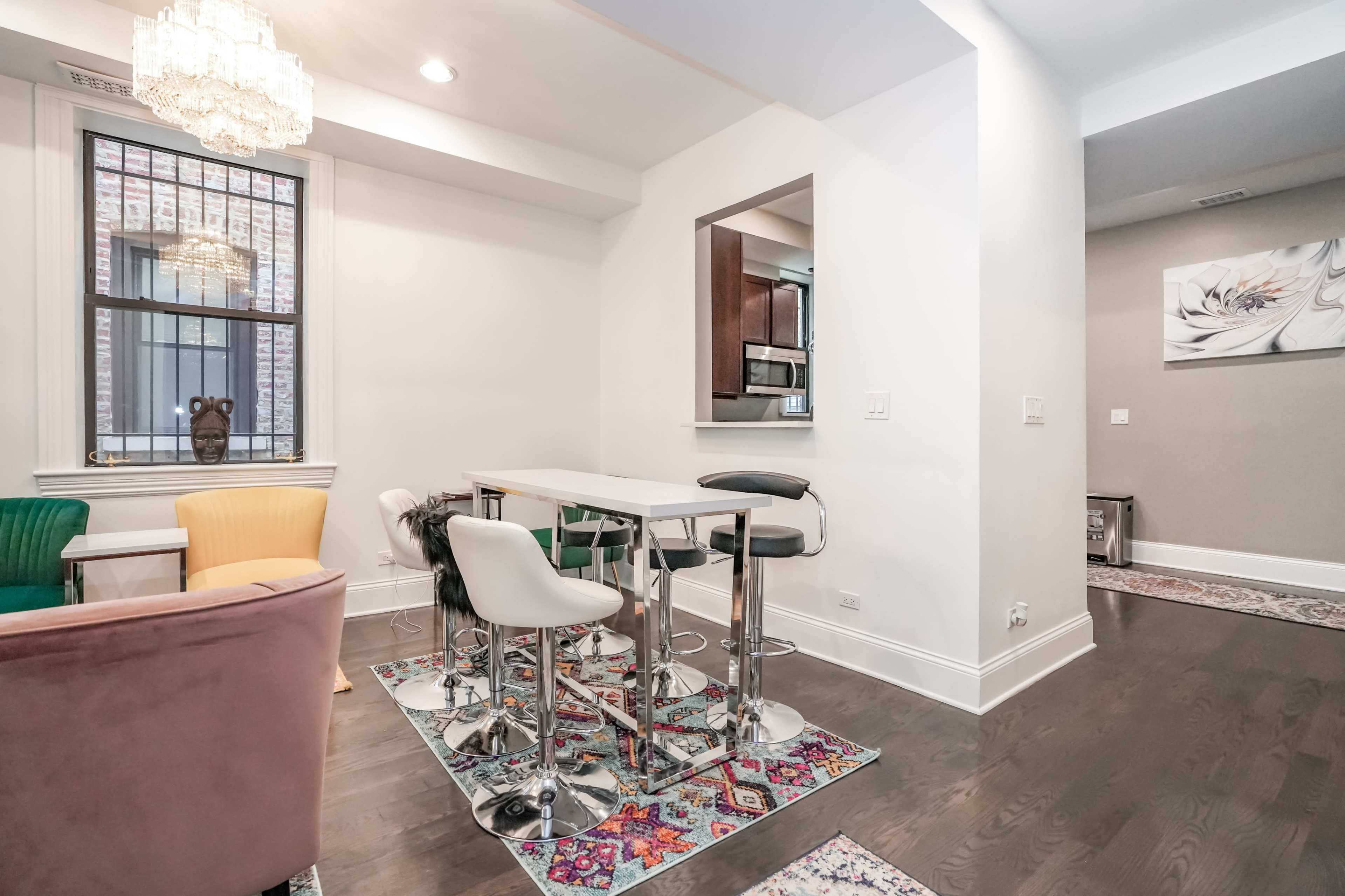 A modern dining area with a table and high stools, surrounded by colorful chairs and a decorative rug on a dark wood floor.
