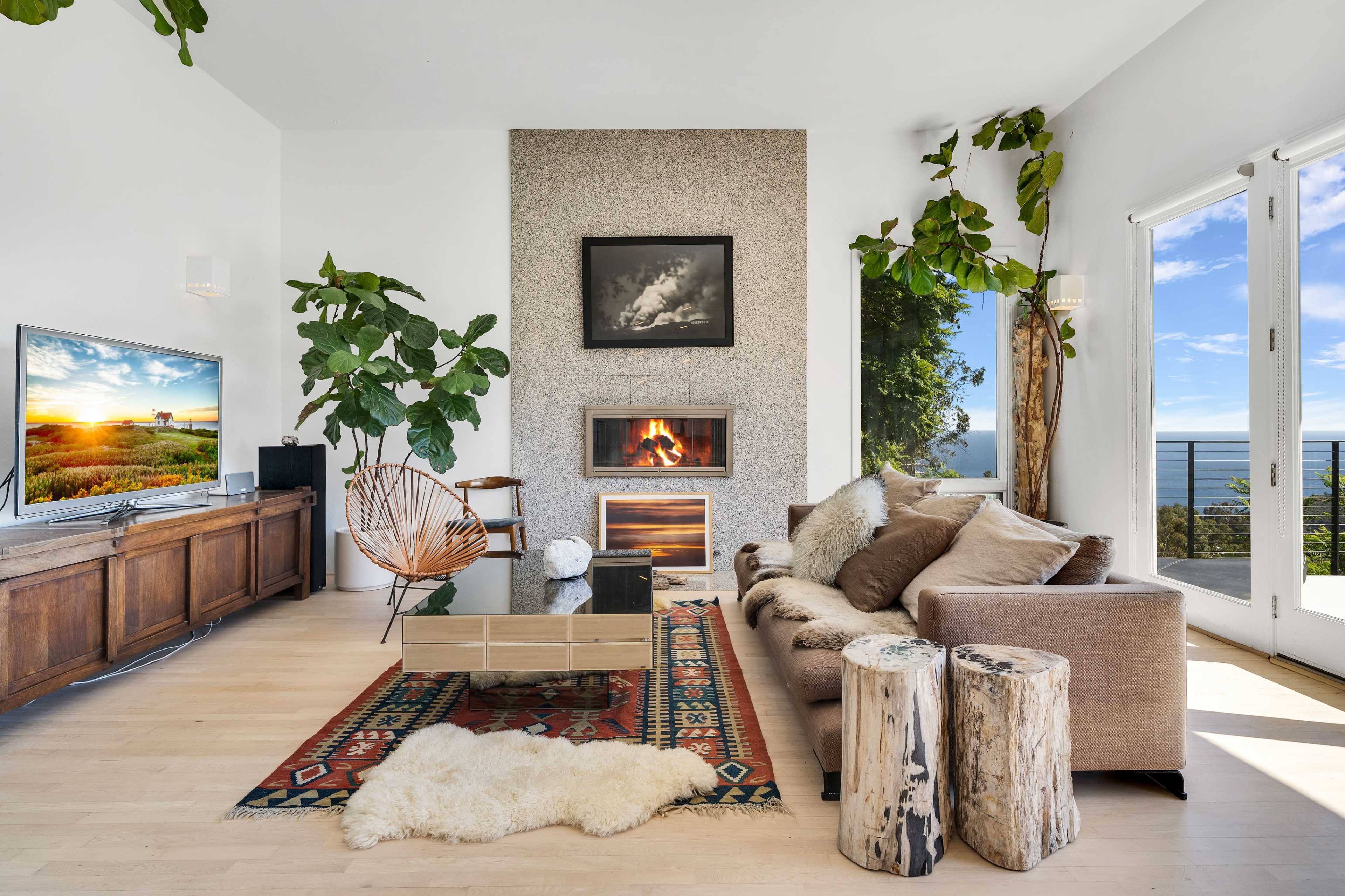 A modern living room features a fireplace, large windows with a view, potted plants, and a combination of wooden and upholstered furniture.