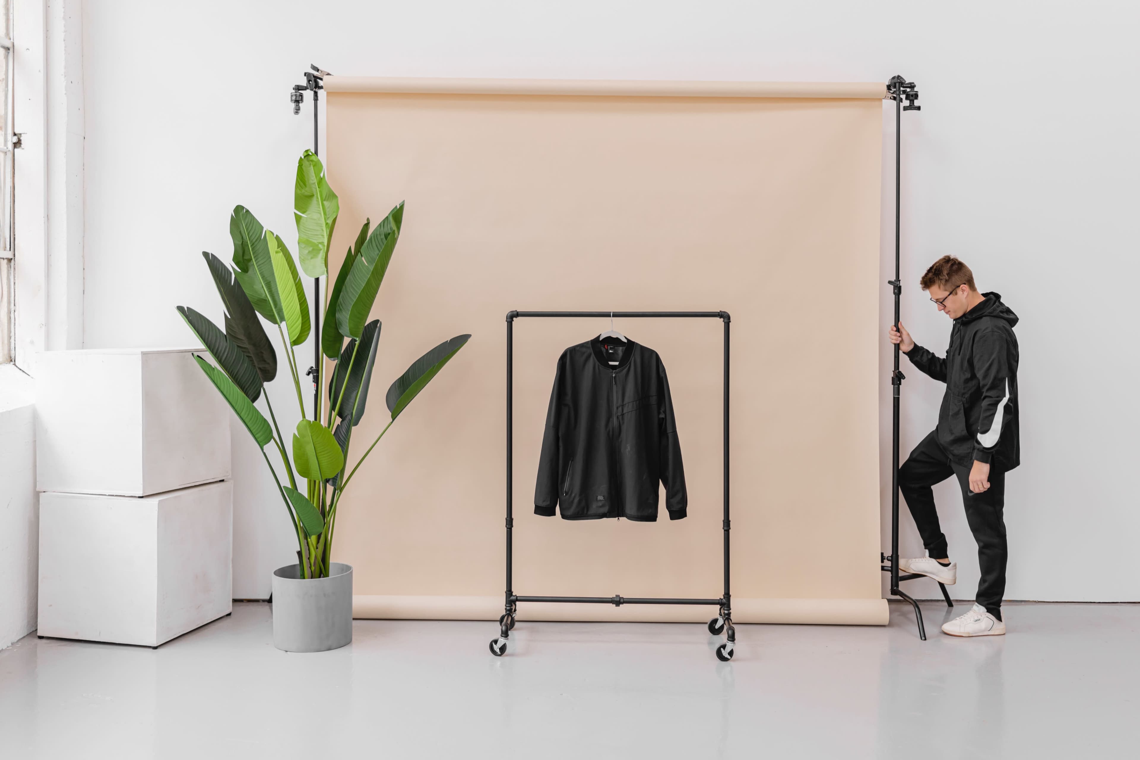 A person stands beside a clothing rack displaying a black jacket against a light backdrop, with a large potted plant nearby.