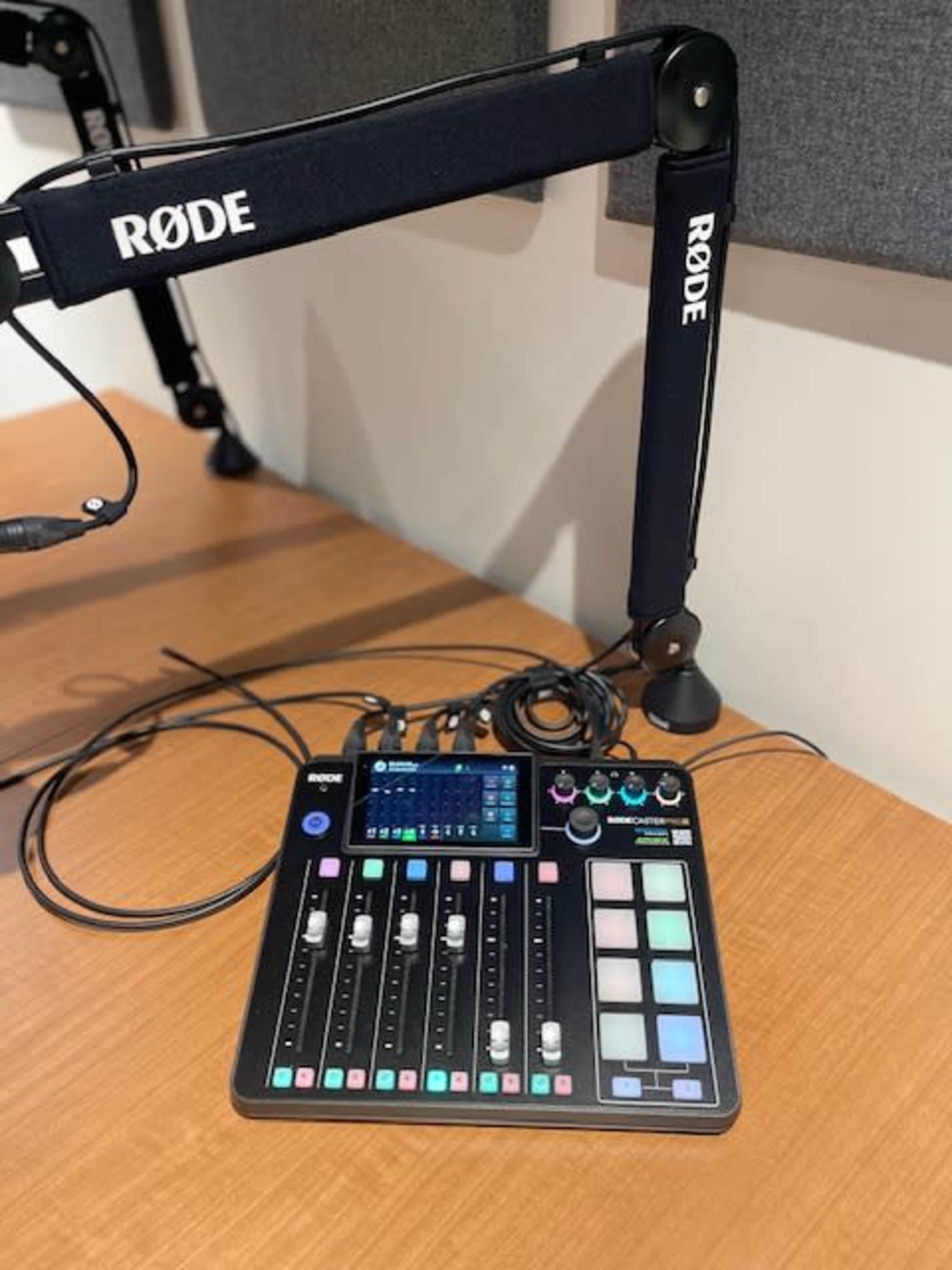 A Rode audio mixing console is positioned on a wooden surface with a microphone arm and cables nearby.