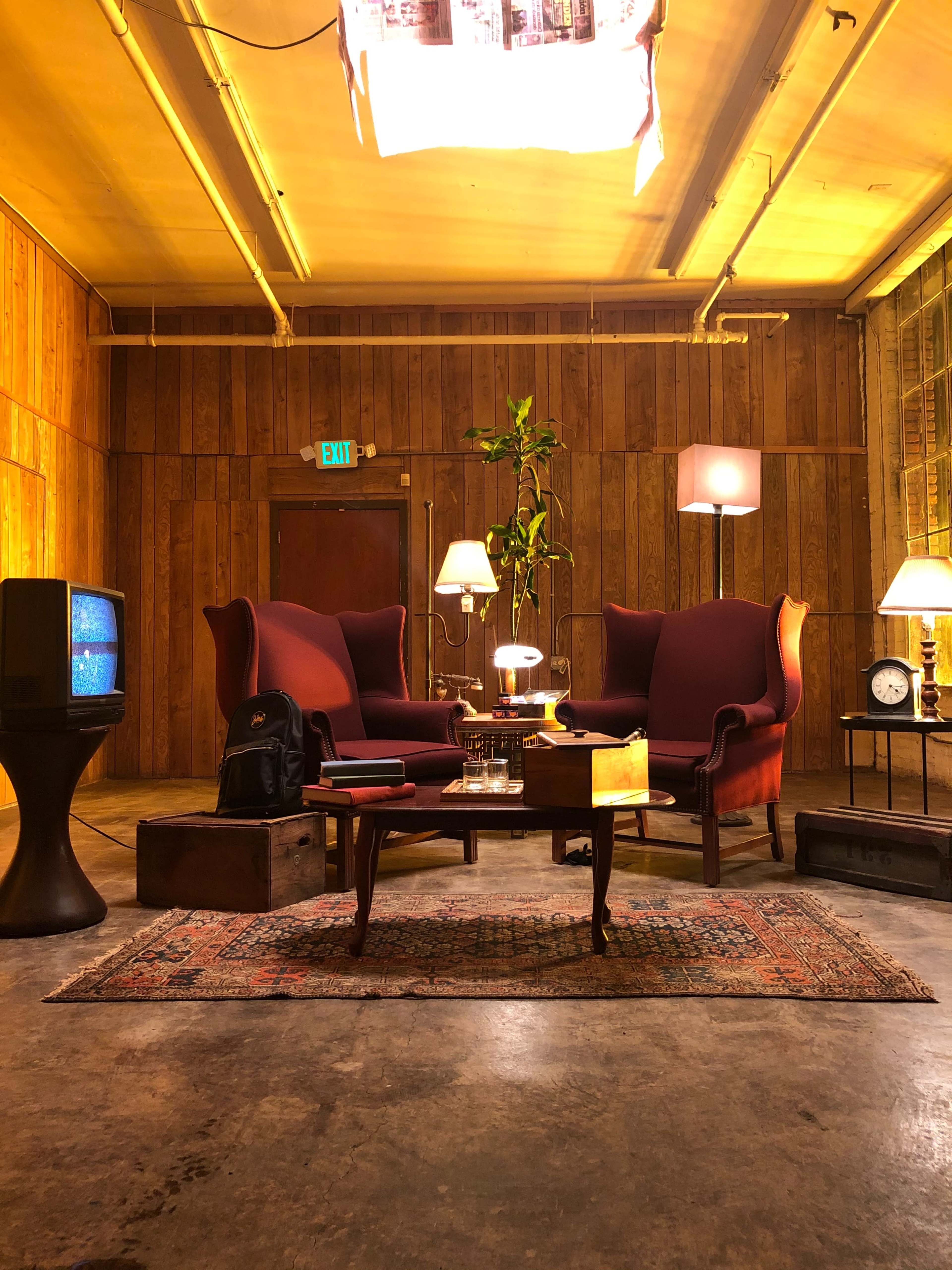 A cozy living room features two armchairs, a coffee table with books and a drink, a small television, and various lamps in a warmly lit wooden interior.