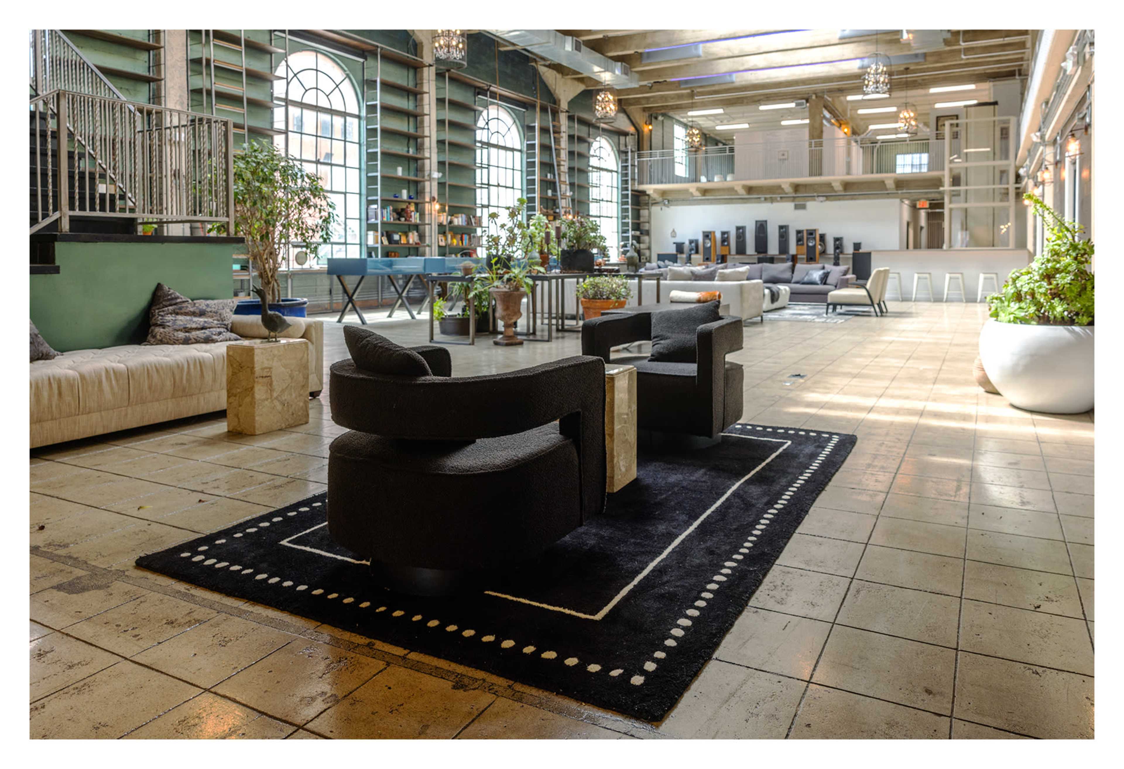 The image shows a spacious interior with large windows, modern furniture, and plants arranged around a central seating area.