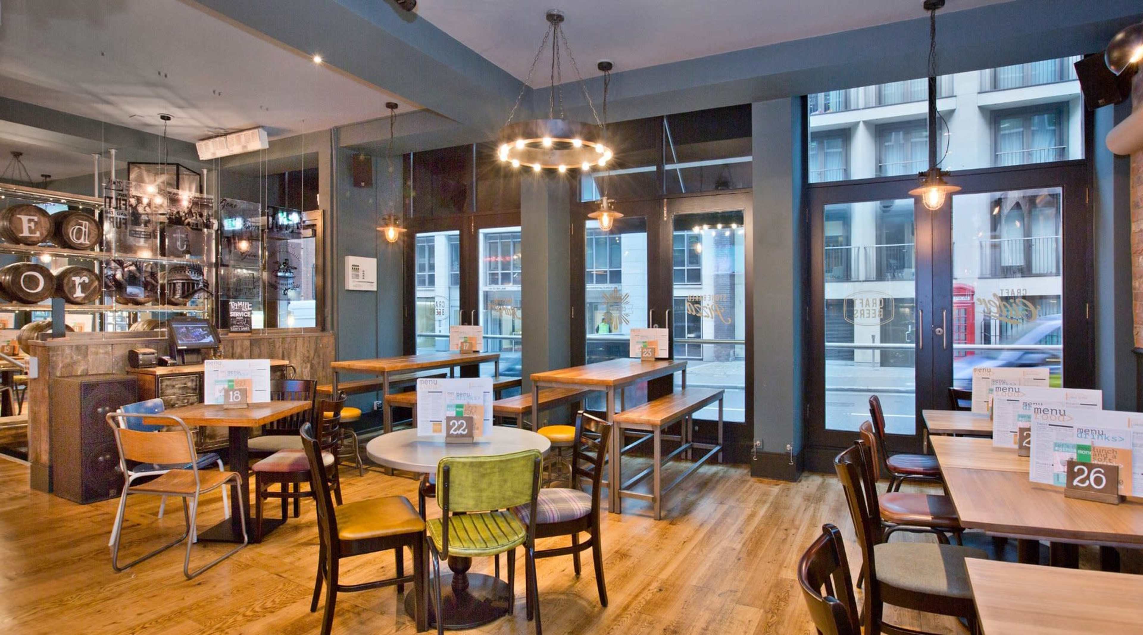 The image shows a modern, well-lit restaurant interior with wooden tables and various seating arrangements, featuring large windows facing the street.