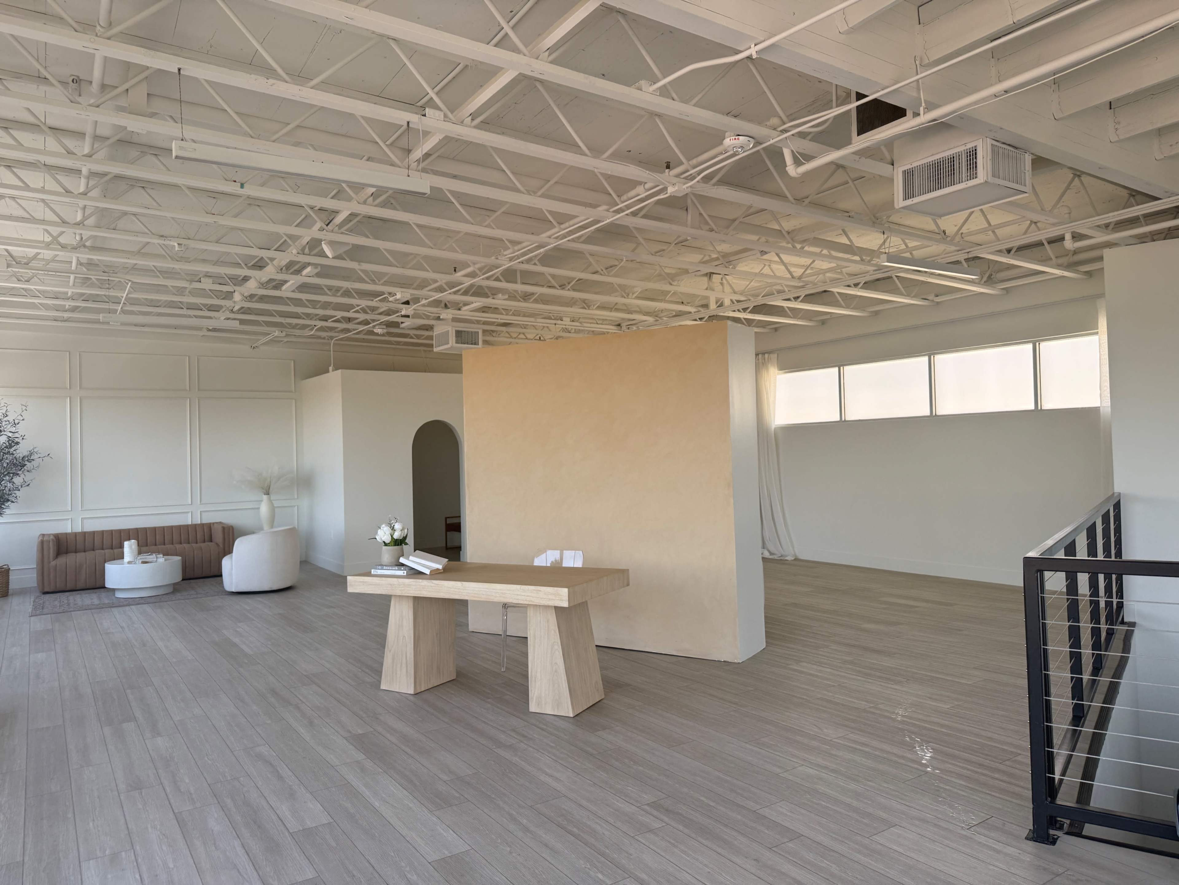 The image shows an empty, modern interior space with wooden flooring, a beige accent wall, and minimal furnishings including a table and seating.