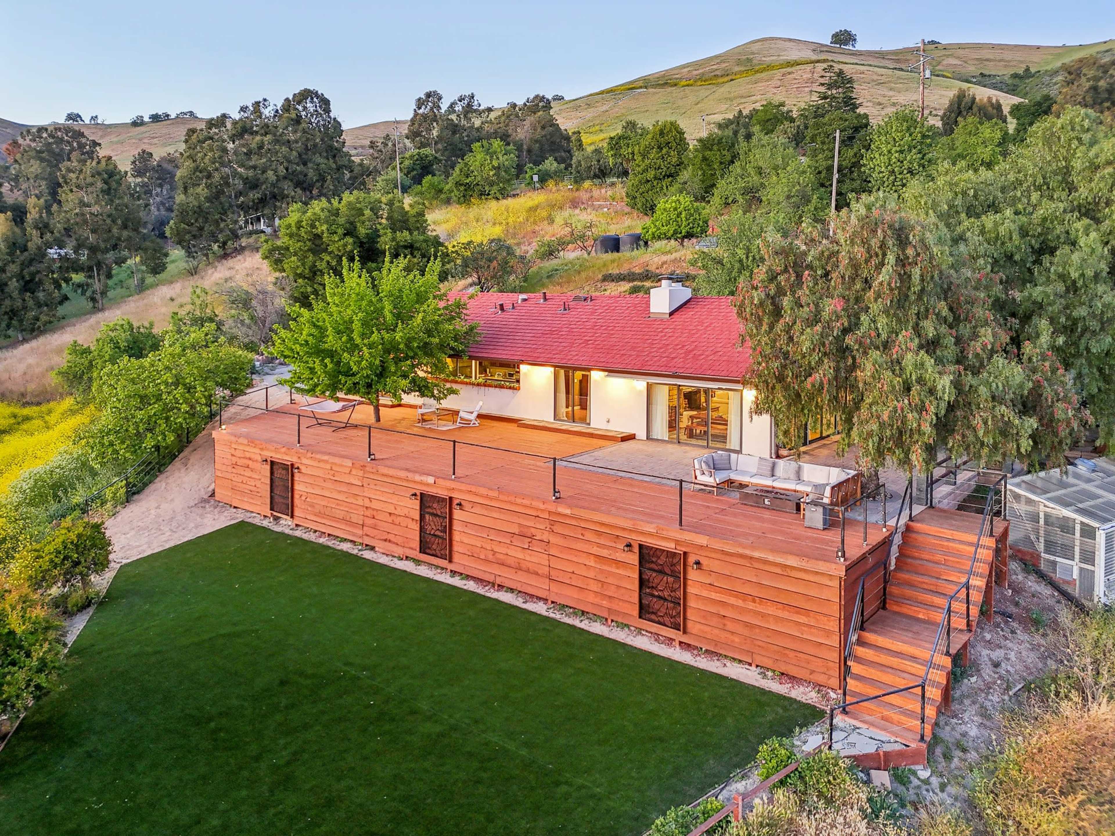 A modern single-story house with a red roof and wooden exterior sits on a hillside surrounded by greenery and a landscaped lawn.
