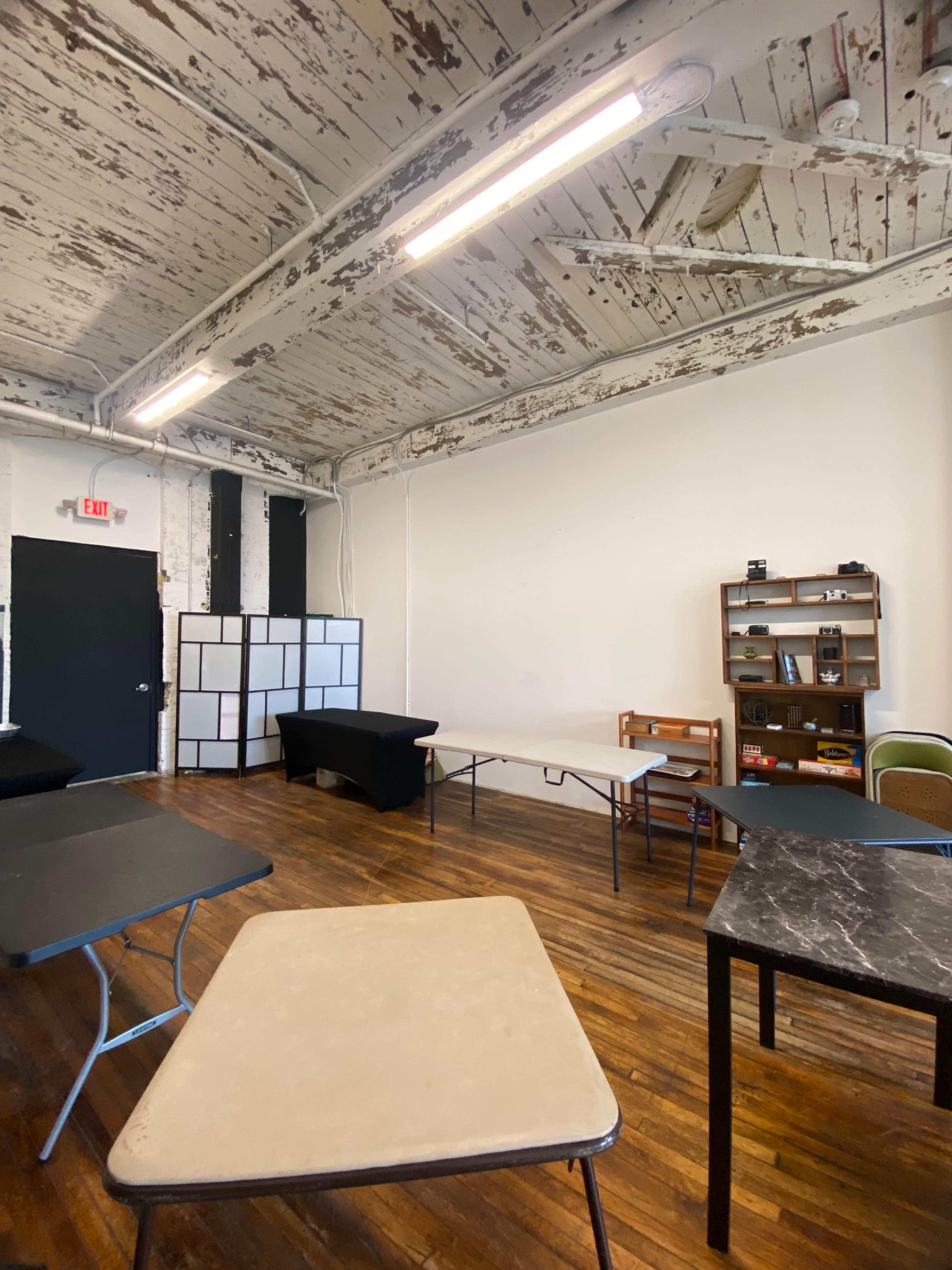 The image shows a sparsely furnished room with wooden floors, several tables of varying shapes and materials, and exposed beams on the ceiling.
