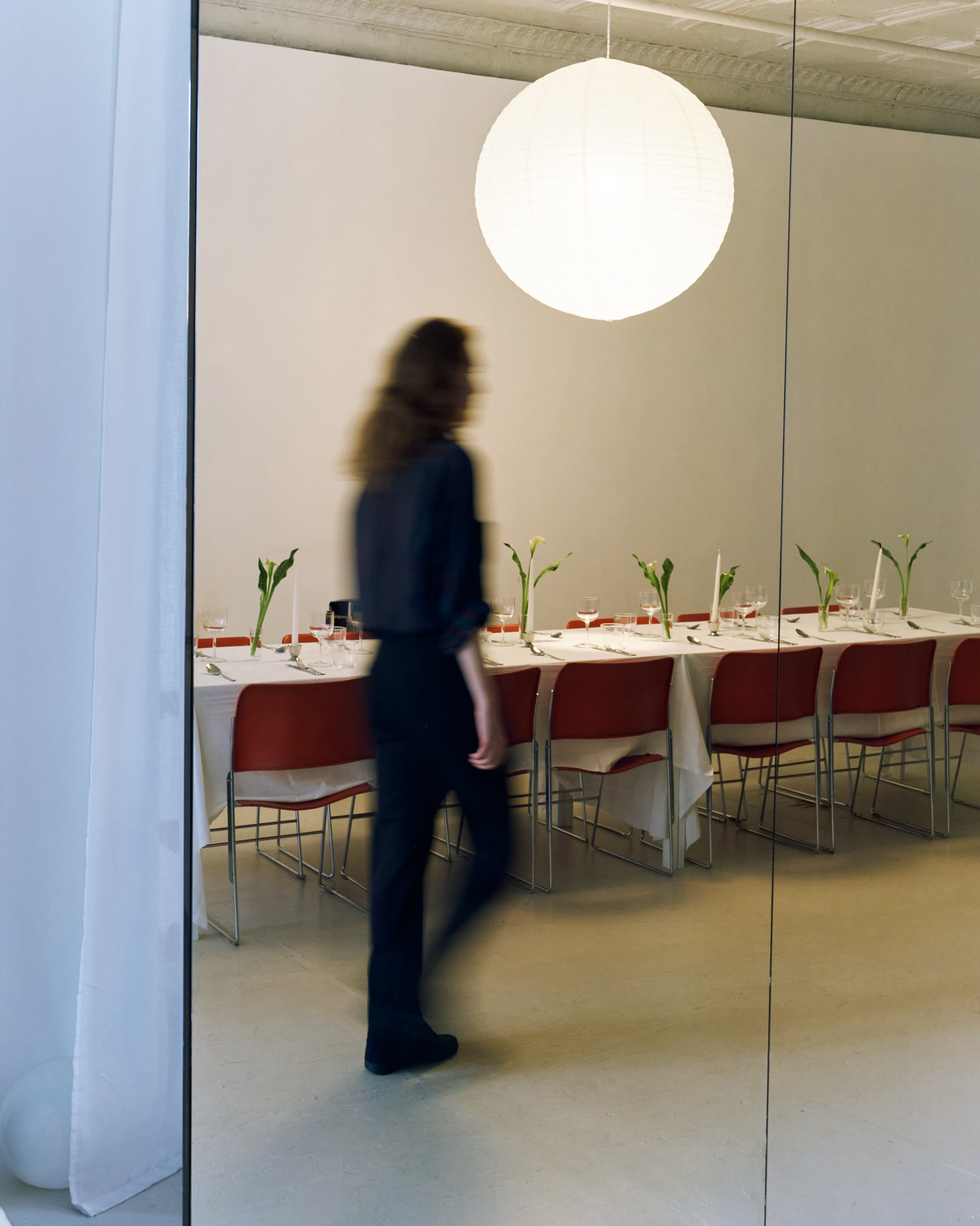 A person walks past a long dining table set for a meal, illuminated by a large hanging paper lantern.