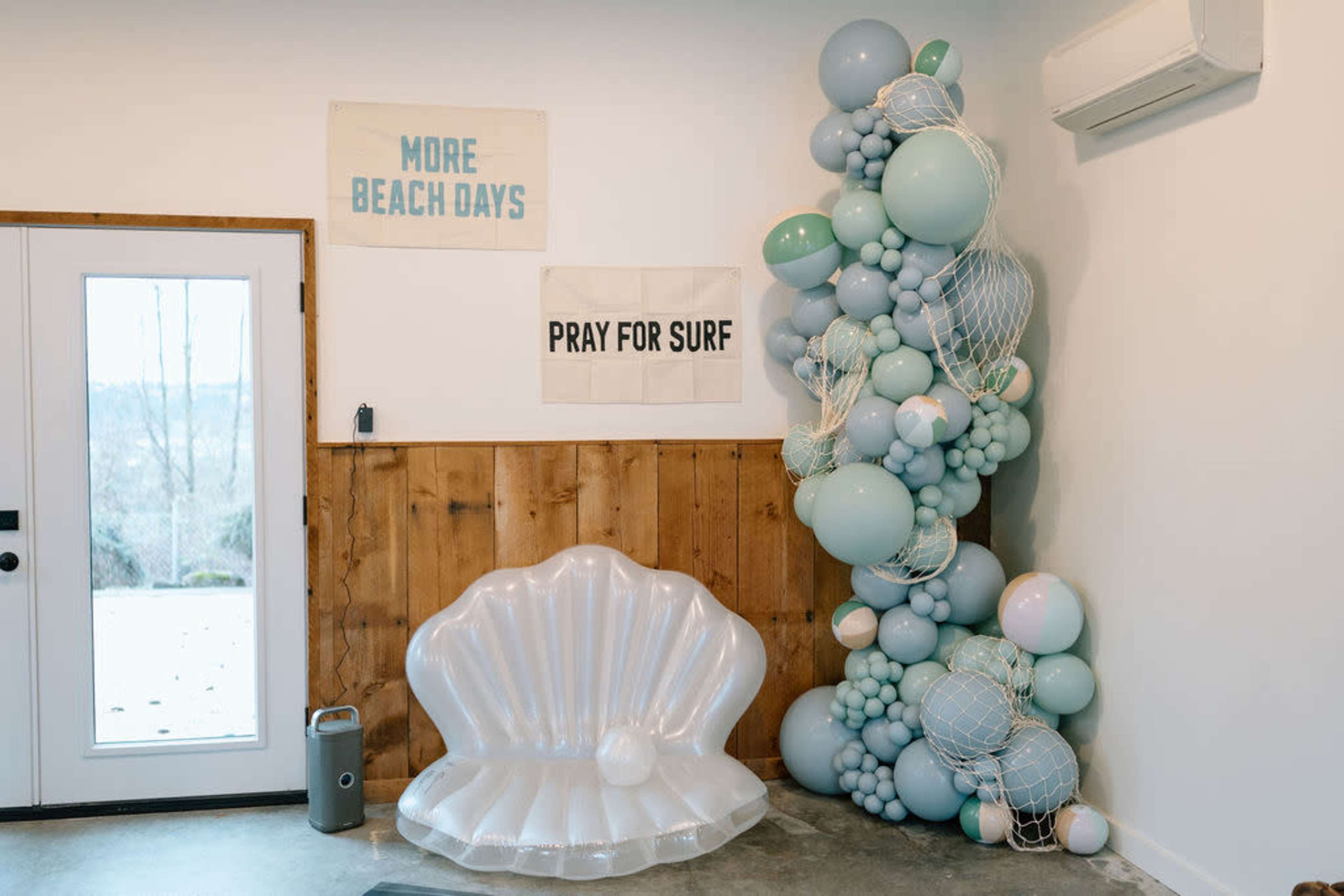 The scene features a decorative corner with a large inflatable clam shell seat, a cluster of pastel-colored balloons arranged artistically, and two signs that read "MORE BEACH DAYS" and "PRAY FOR SURF."