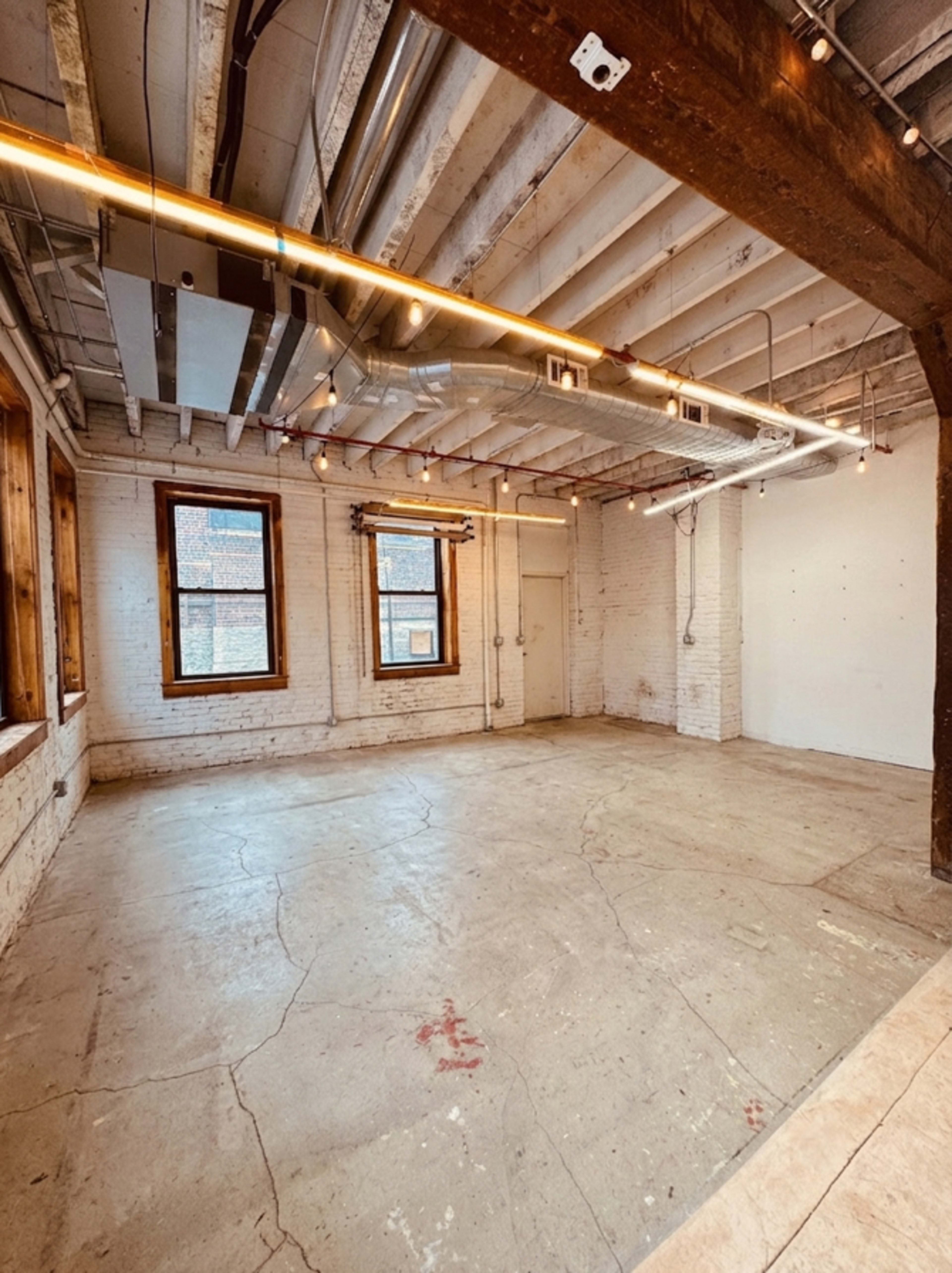 The image shows an empty industrial-style room with exposed brick walls, wooden window frames, and overhead lighting.