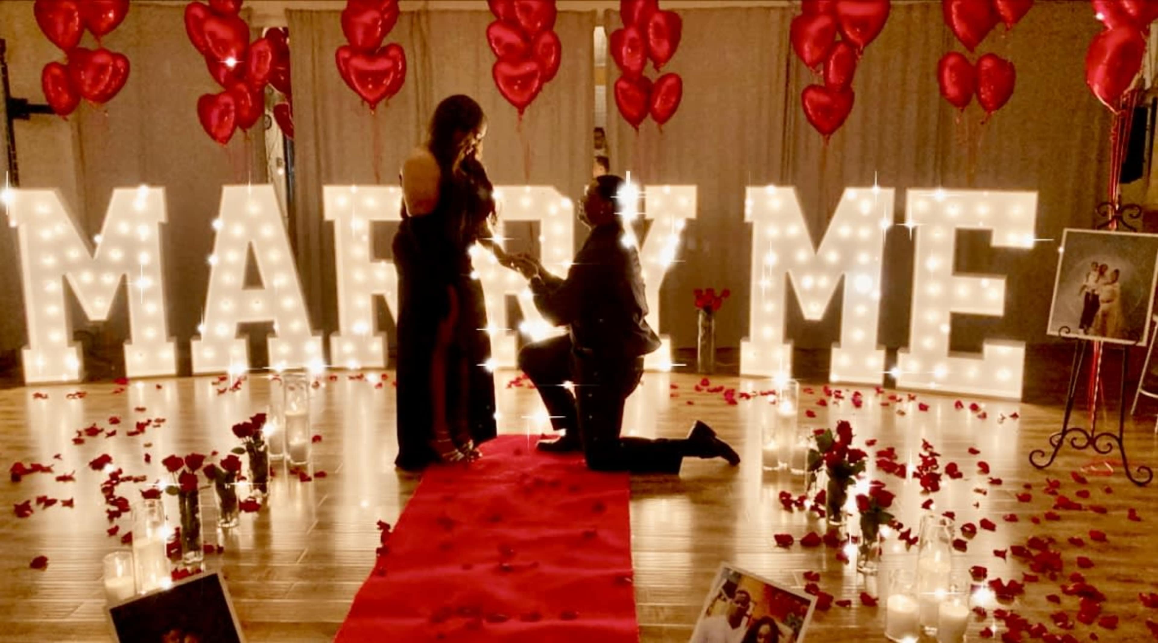 A man kneels on a red carpet surrounded by rose petals and candlelight, proposing to a woman in front of illuminated letters that spell "MARRY ME," with heart-shaped balloons above.