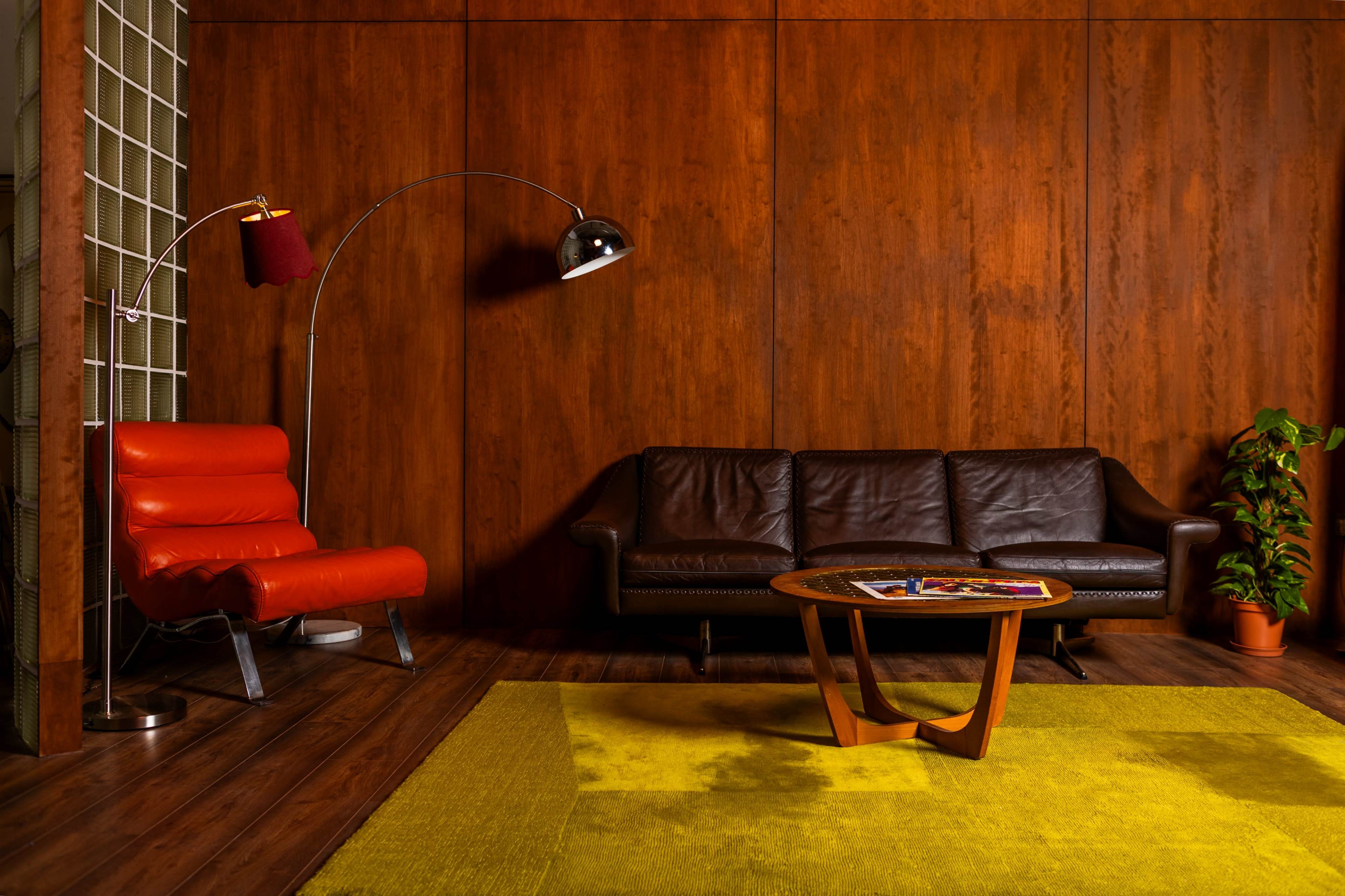 The image features a modern, minimalist living room with a wooden wall, a red and black sofa, a curved floor lamp, a round coffee table, and a potted plant.