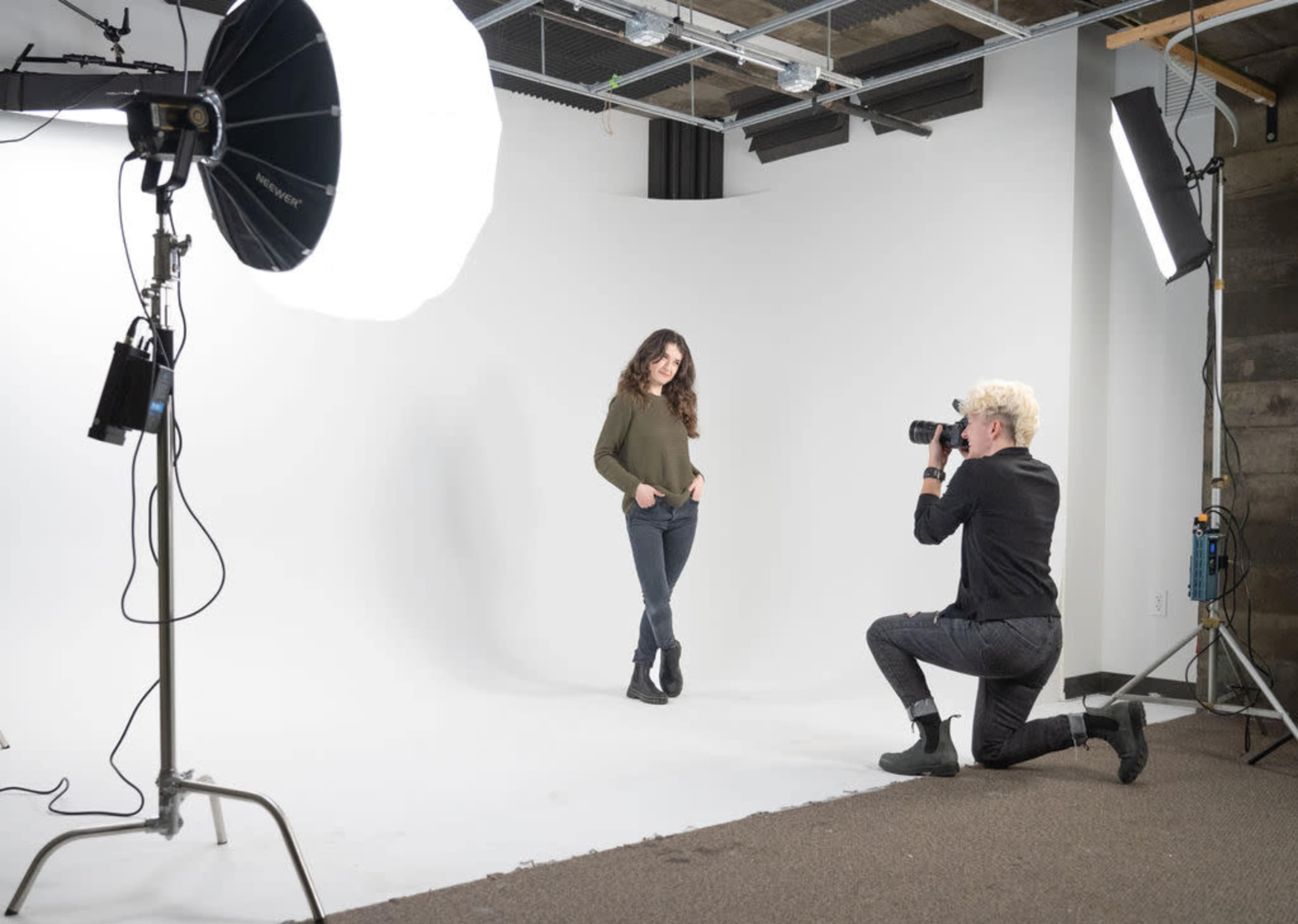 A photographer kneels while taking pictures of a model in a simple studio setup with a white backdrop and lighting equipment.