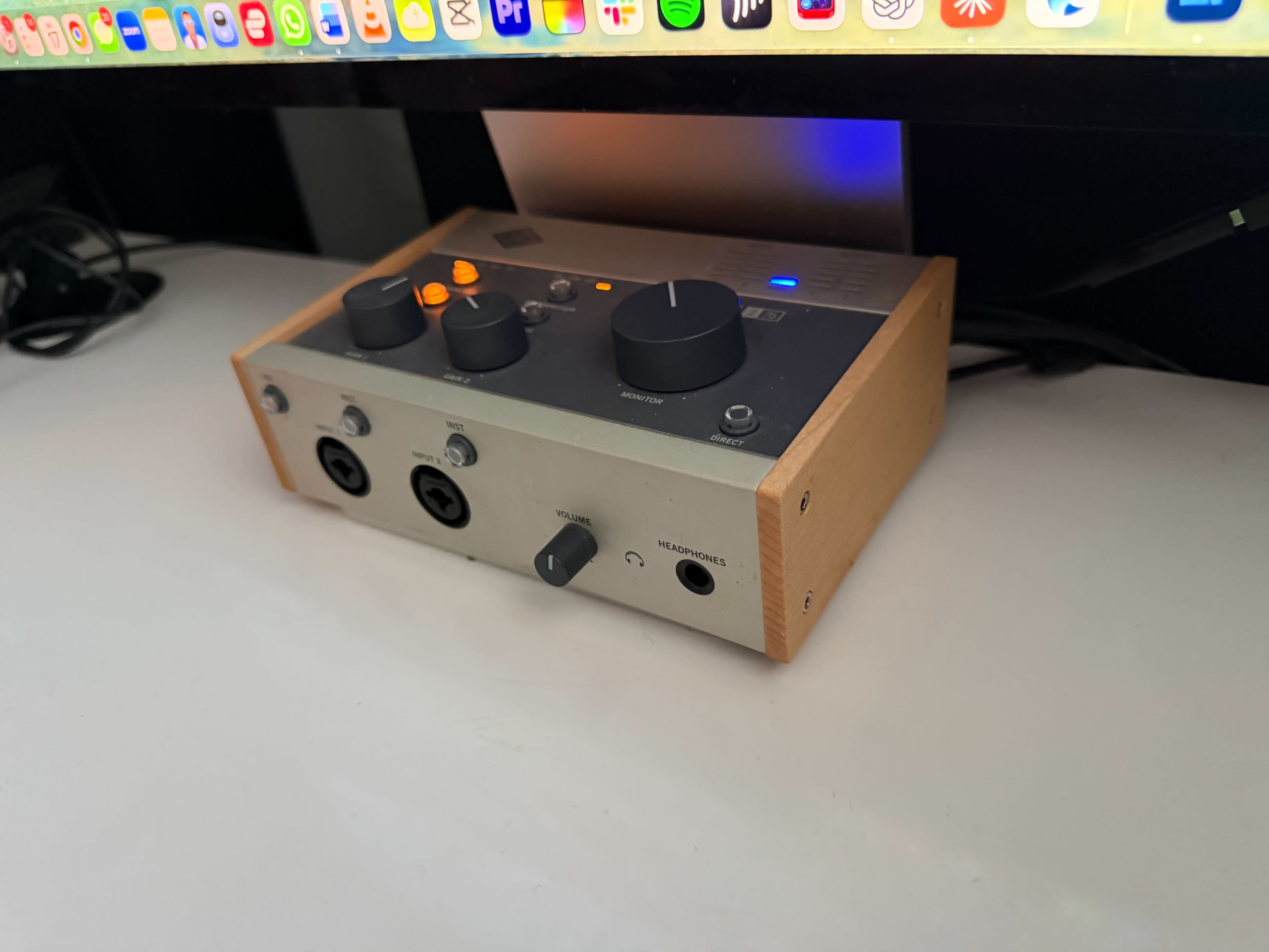 A compact audio interface with various knobs and input/output ports is positioned on a white desk beneath a computer monitor.