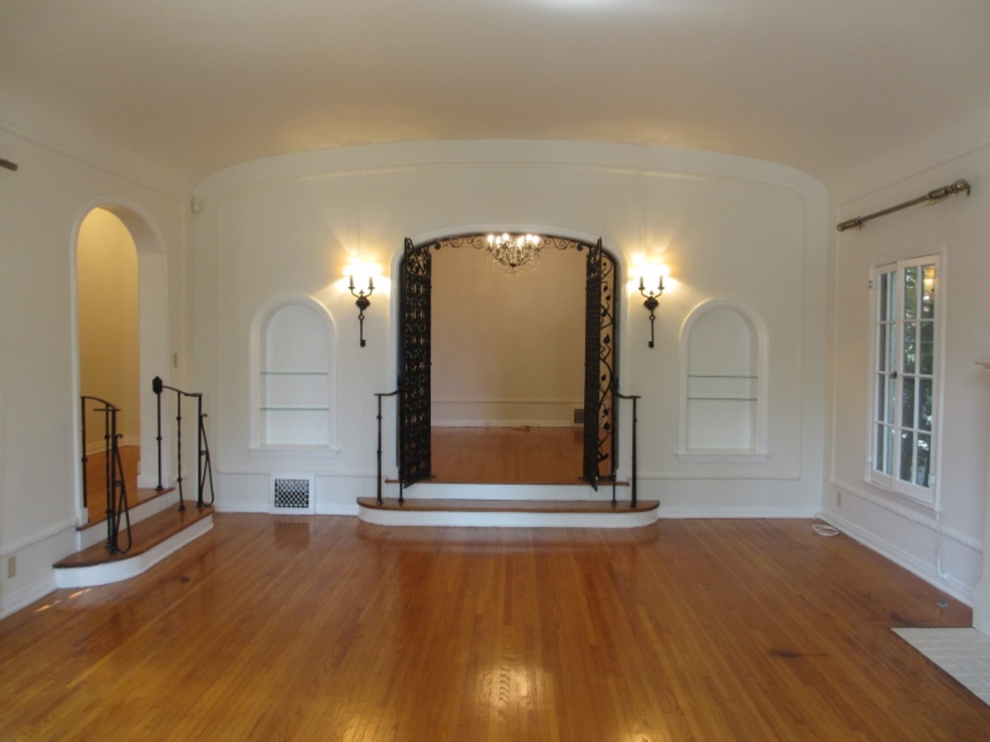 The image shows a spacious room featuring an archway with decorative ironwork, two wall sconces, and hardwood flooring.