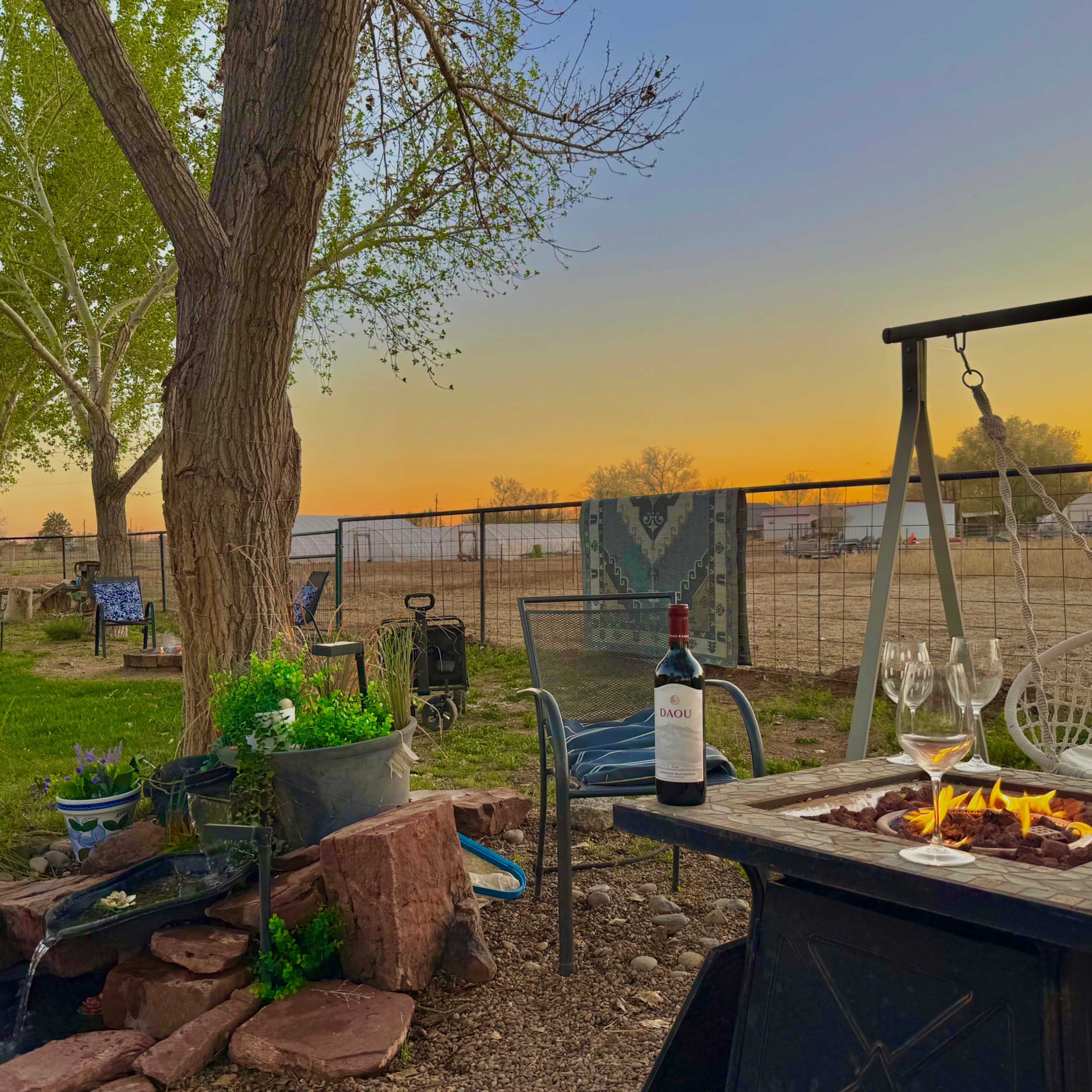 A bottle of wine and two glasses sit on a table beside a fire pit, with a sunset and grassy landscape visible in the background.