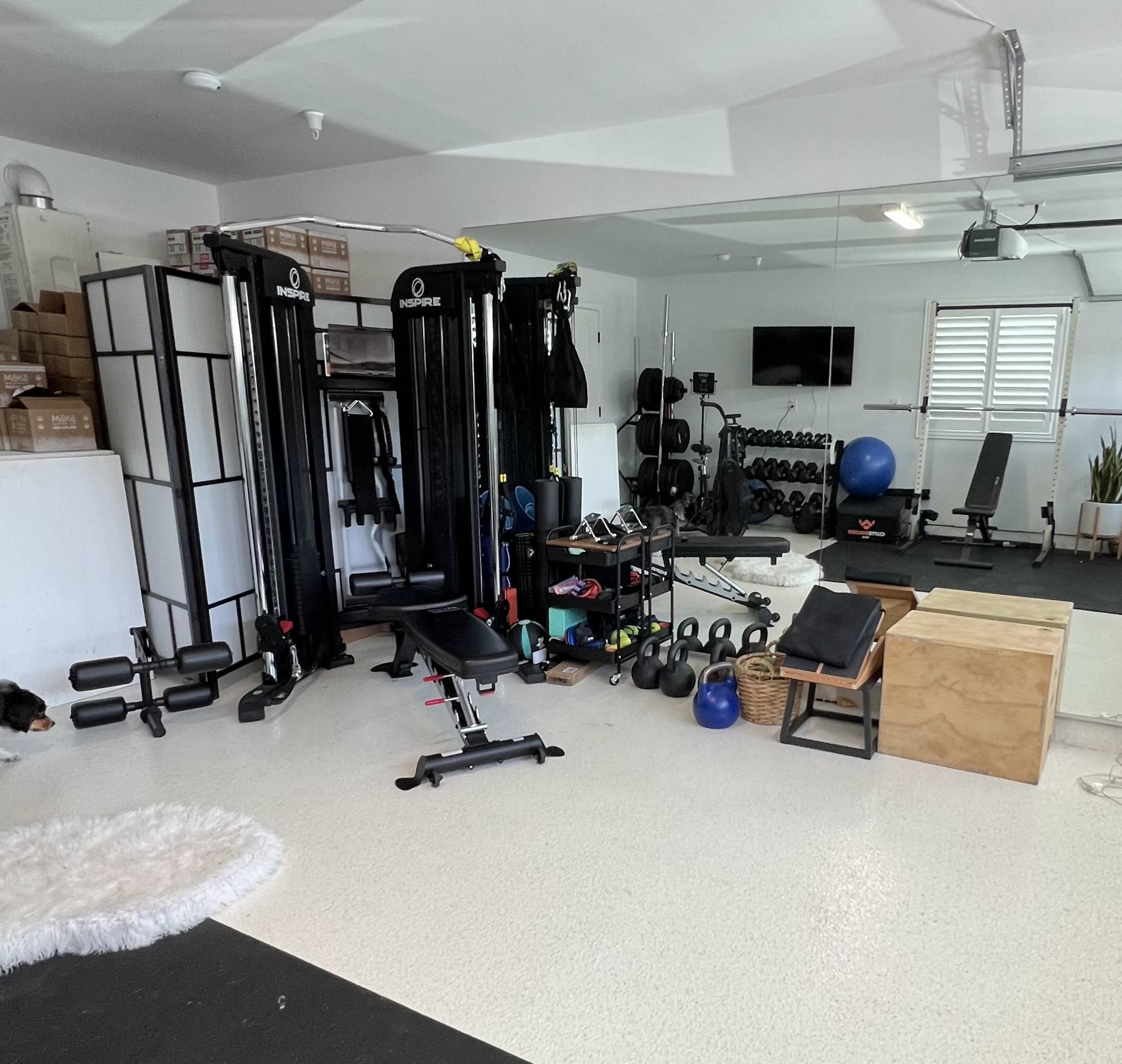 A home gym setup with various exercise equipment, including weight machines, dumbbells, a stability ball, and a wooden box, all arranged in a spacious garage.