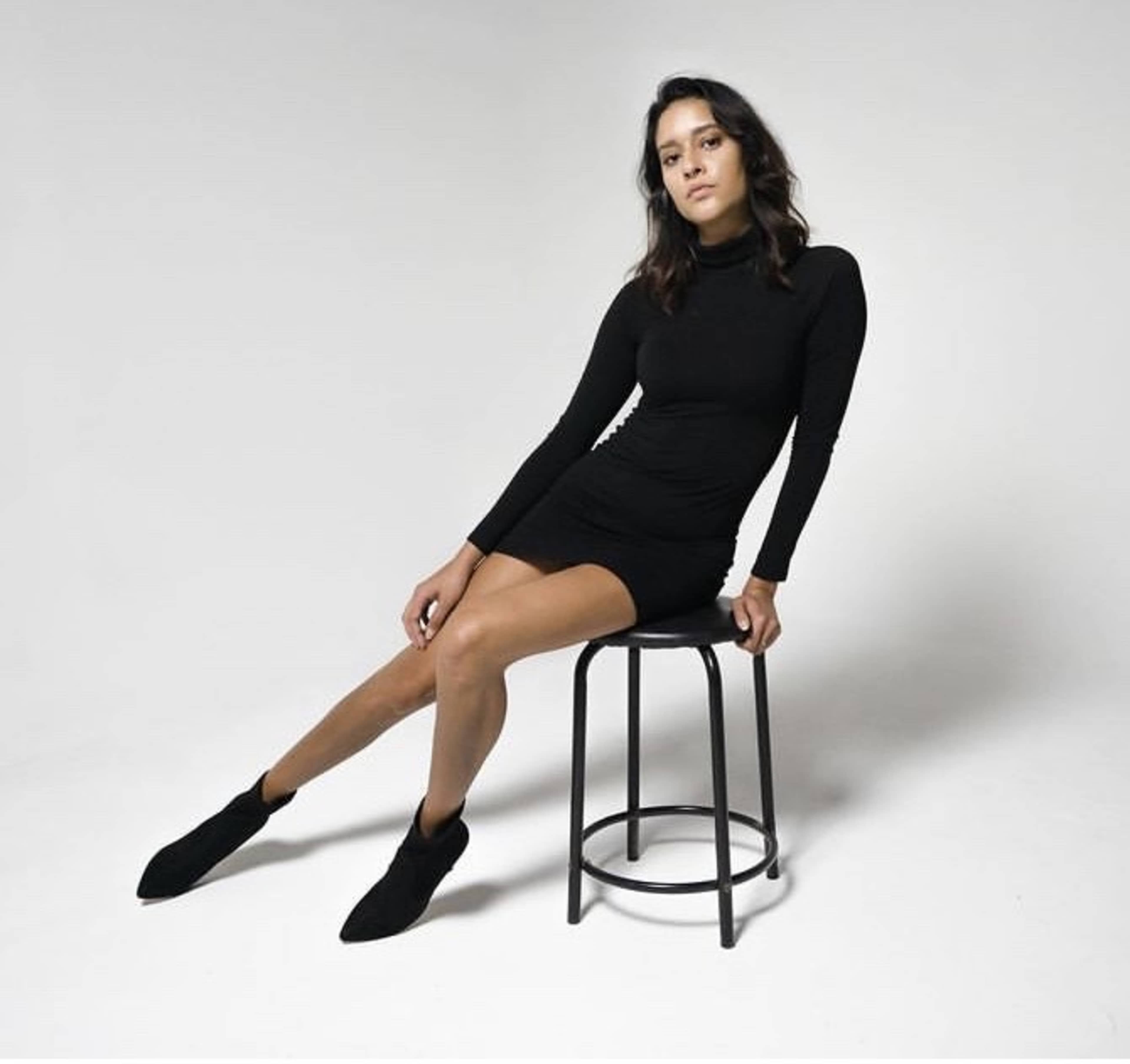 A woman in a black long-sleeve dress sits on a black stool with one leg extended.