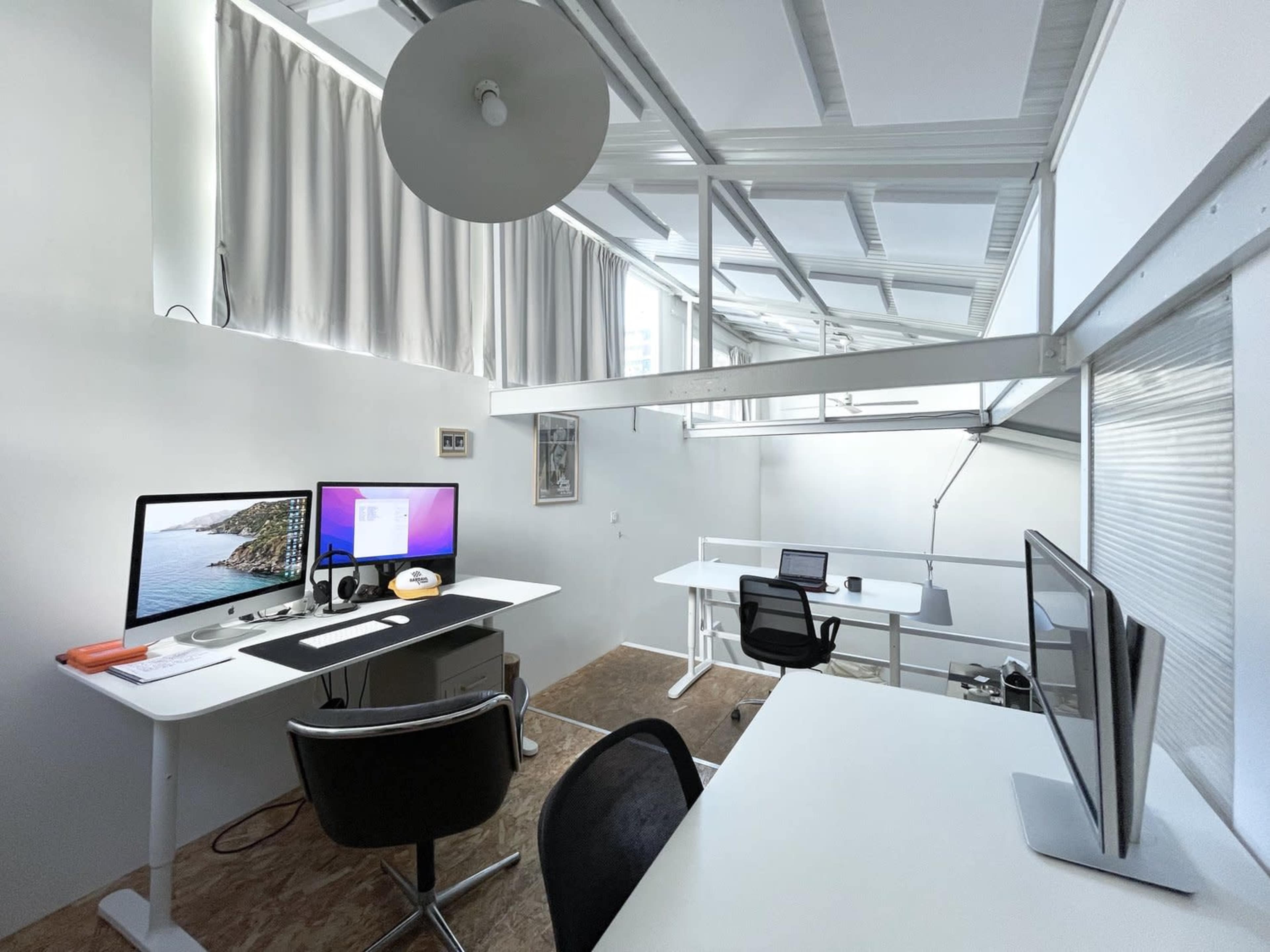 A modern workspace featuring two computer desks with monitors, a chair, and large windows allowing natural light.