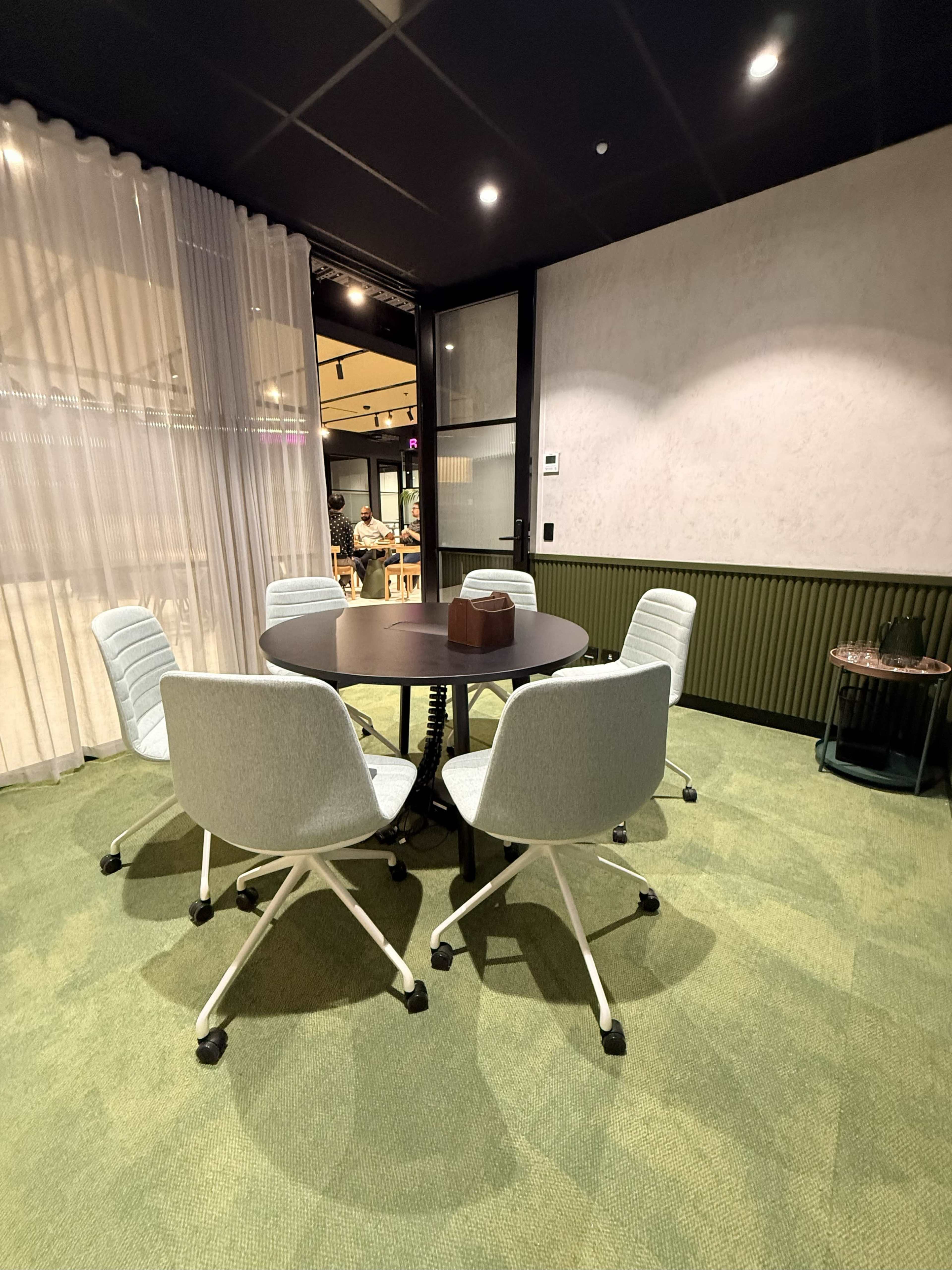 A round table surrounded by four light blue chairs is situated in a modern office meeting room with green carpet and partial curtains.