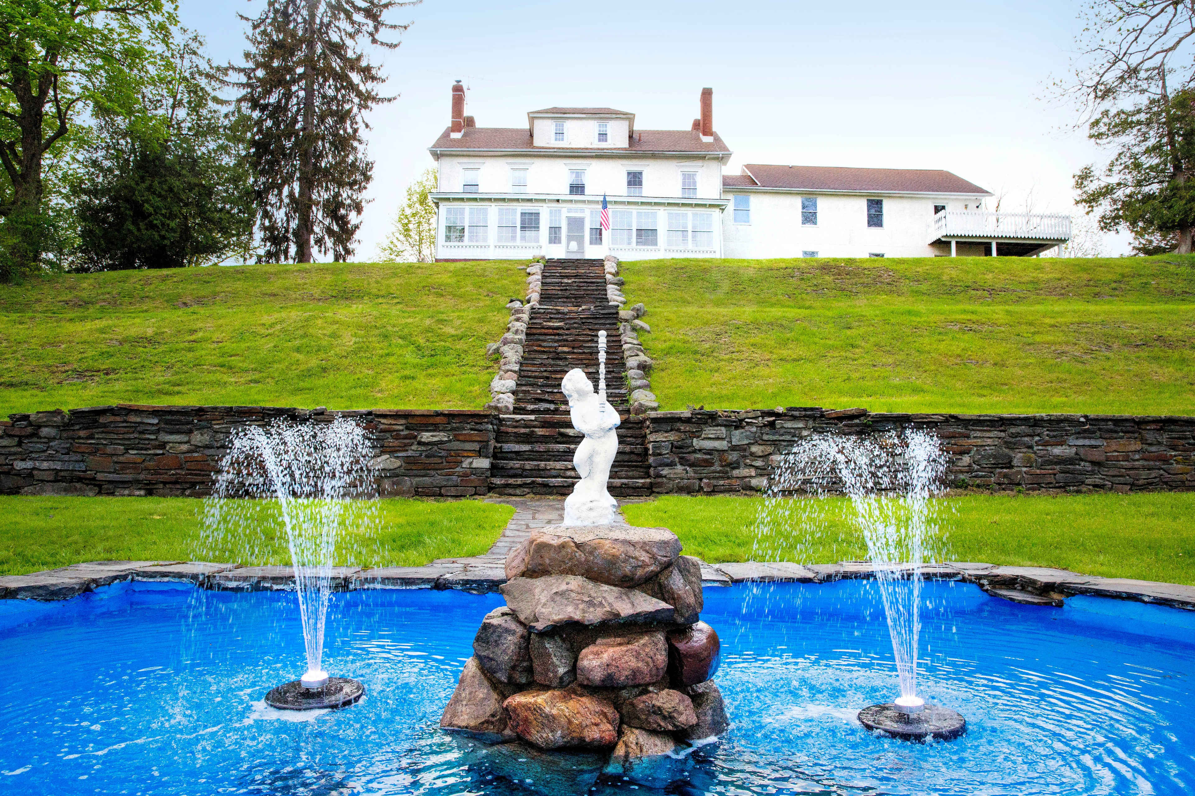 A large white house on a hill, with a stone fountain and blue water in the foreground, and a pathway leading up to the house.
