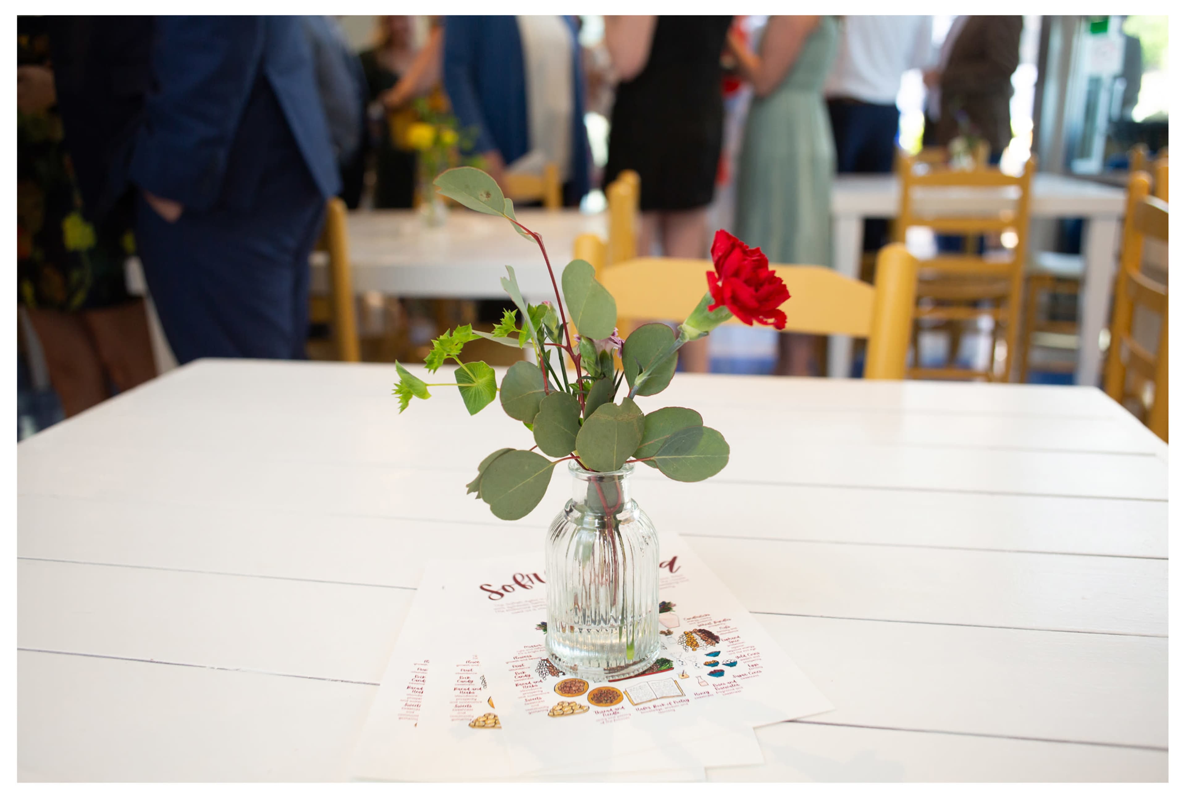 A small glass vase with a red flower and green leaves sits on a white table covered with a menu.