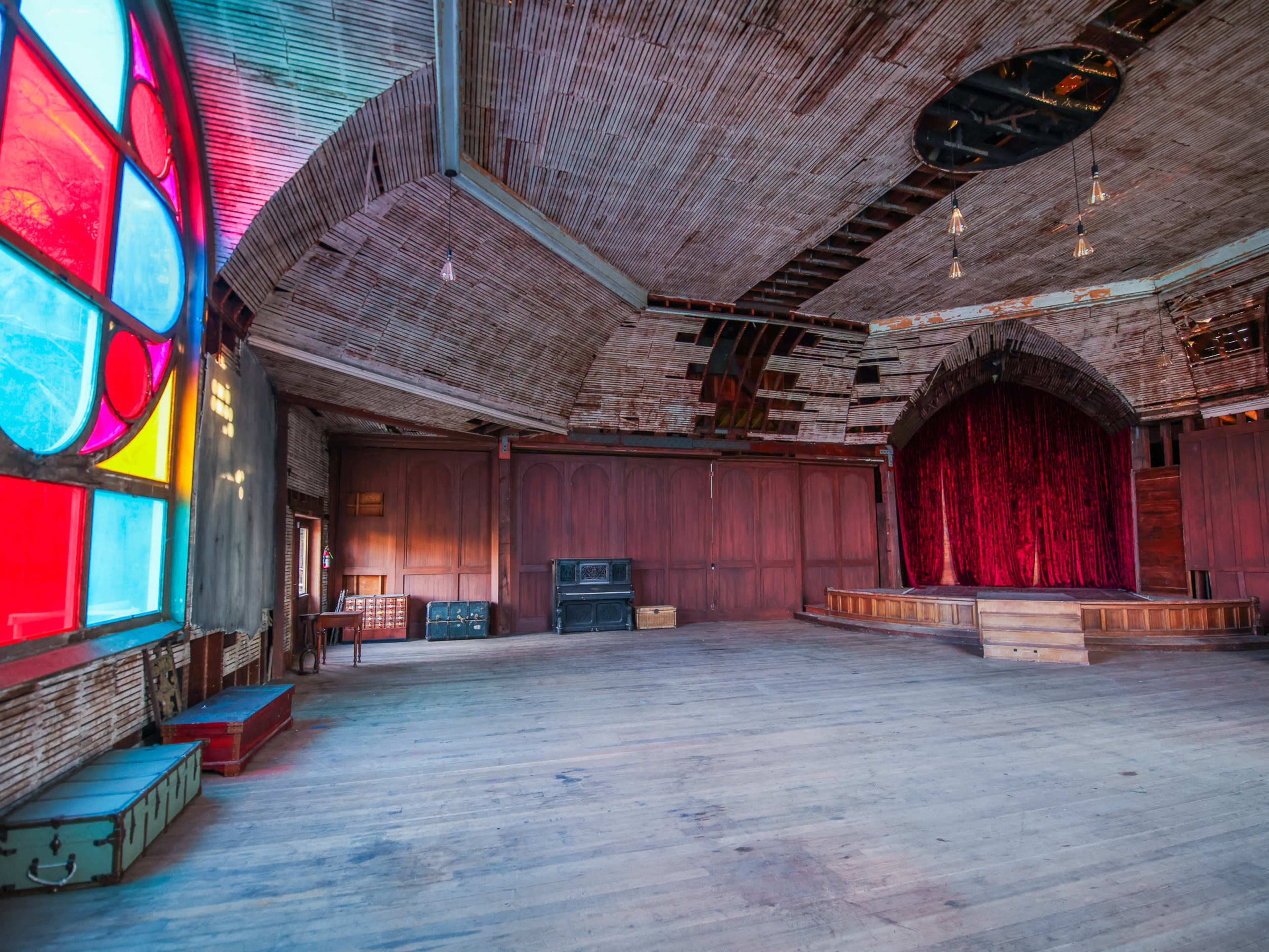 The image shows an empty, rustic hall with a large stained glass window on one side and a red velvet curtain backdrop on a wooden stage.