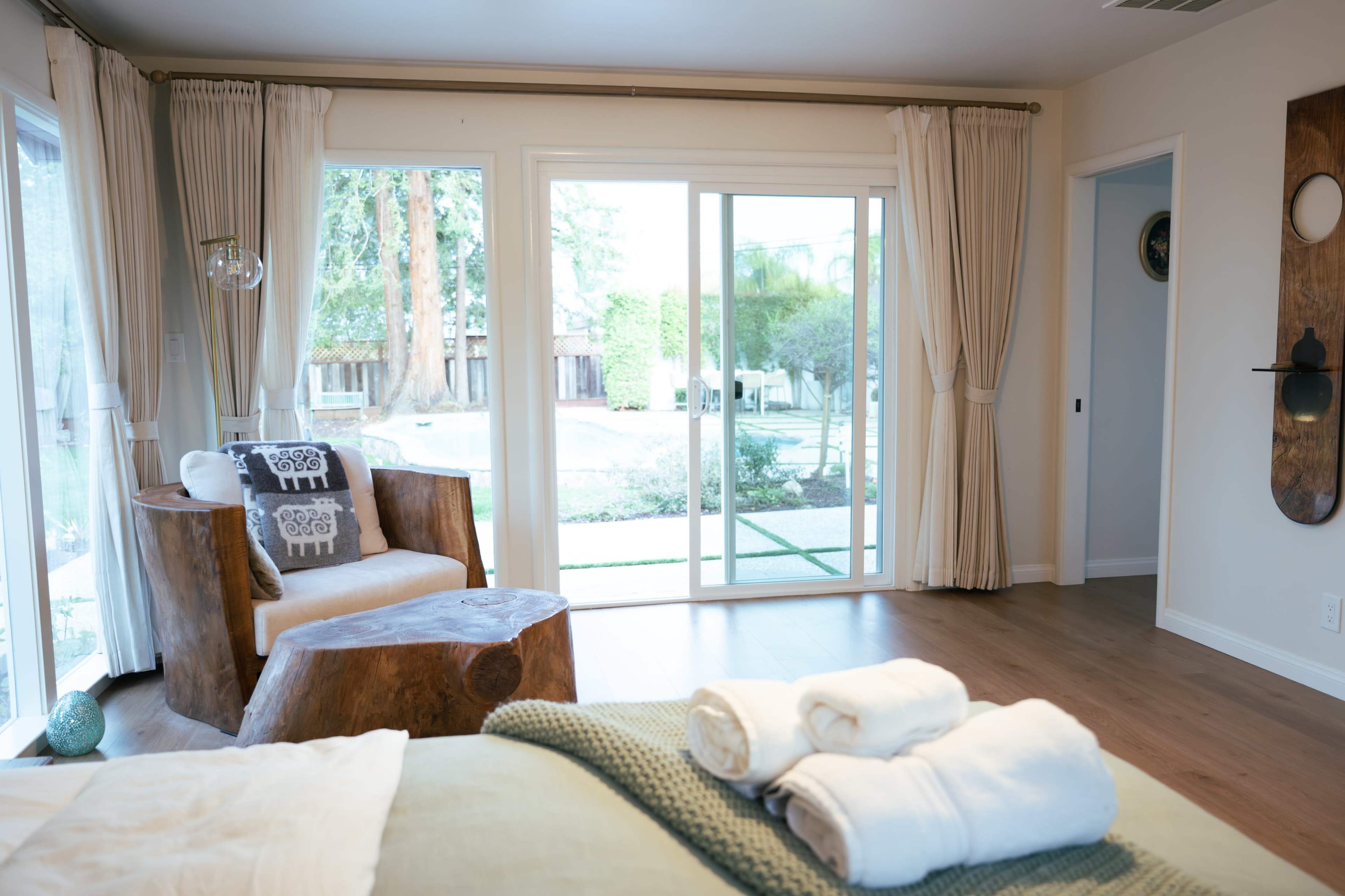 The room features a wooden chair and a small table, with large windows allowing natural light to illuminate a view of a garden.