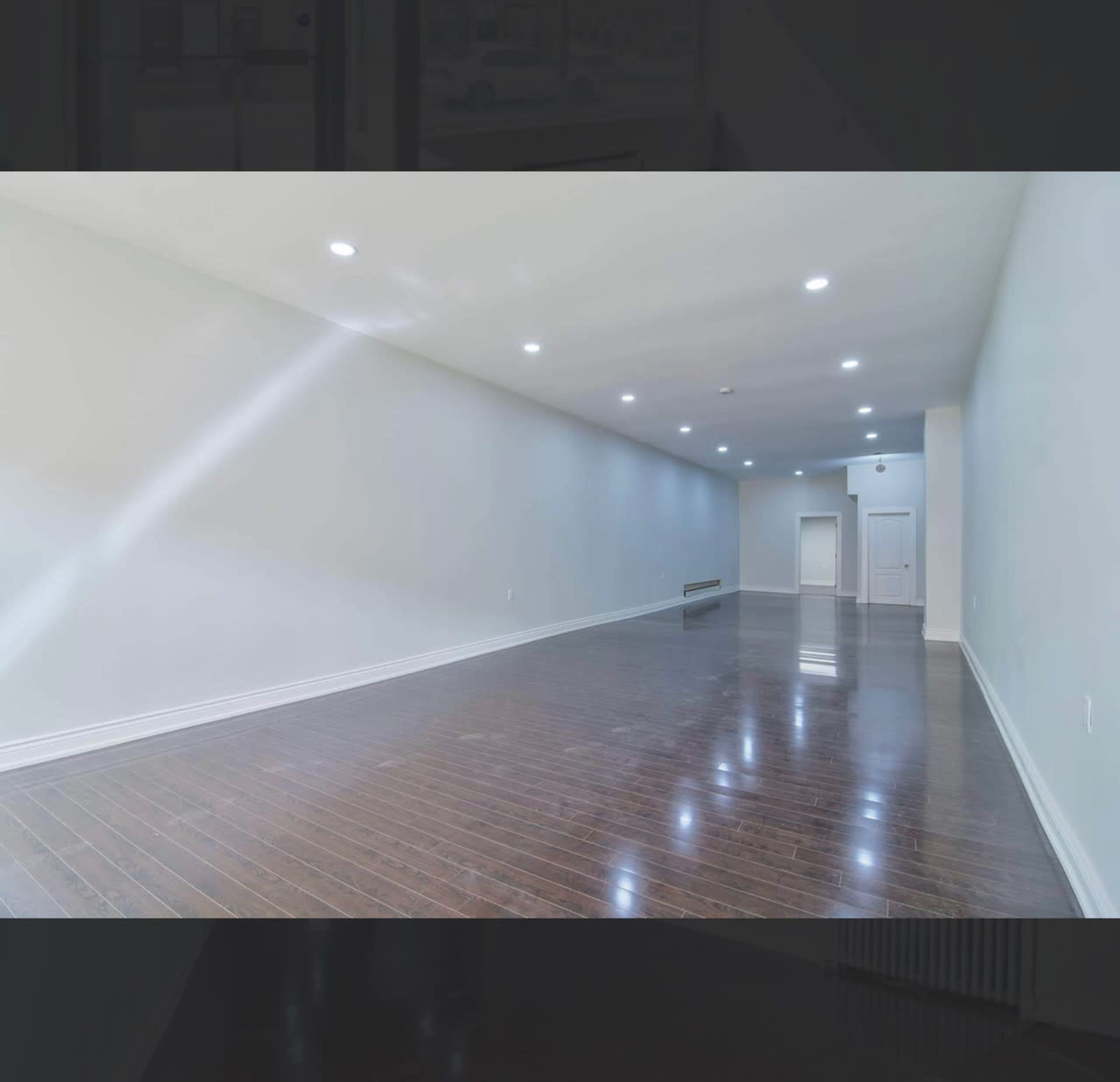 The image shows a spacious room with hardwood flooring and white walls, illuminated by ceiling lights.