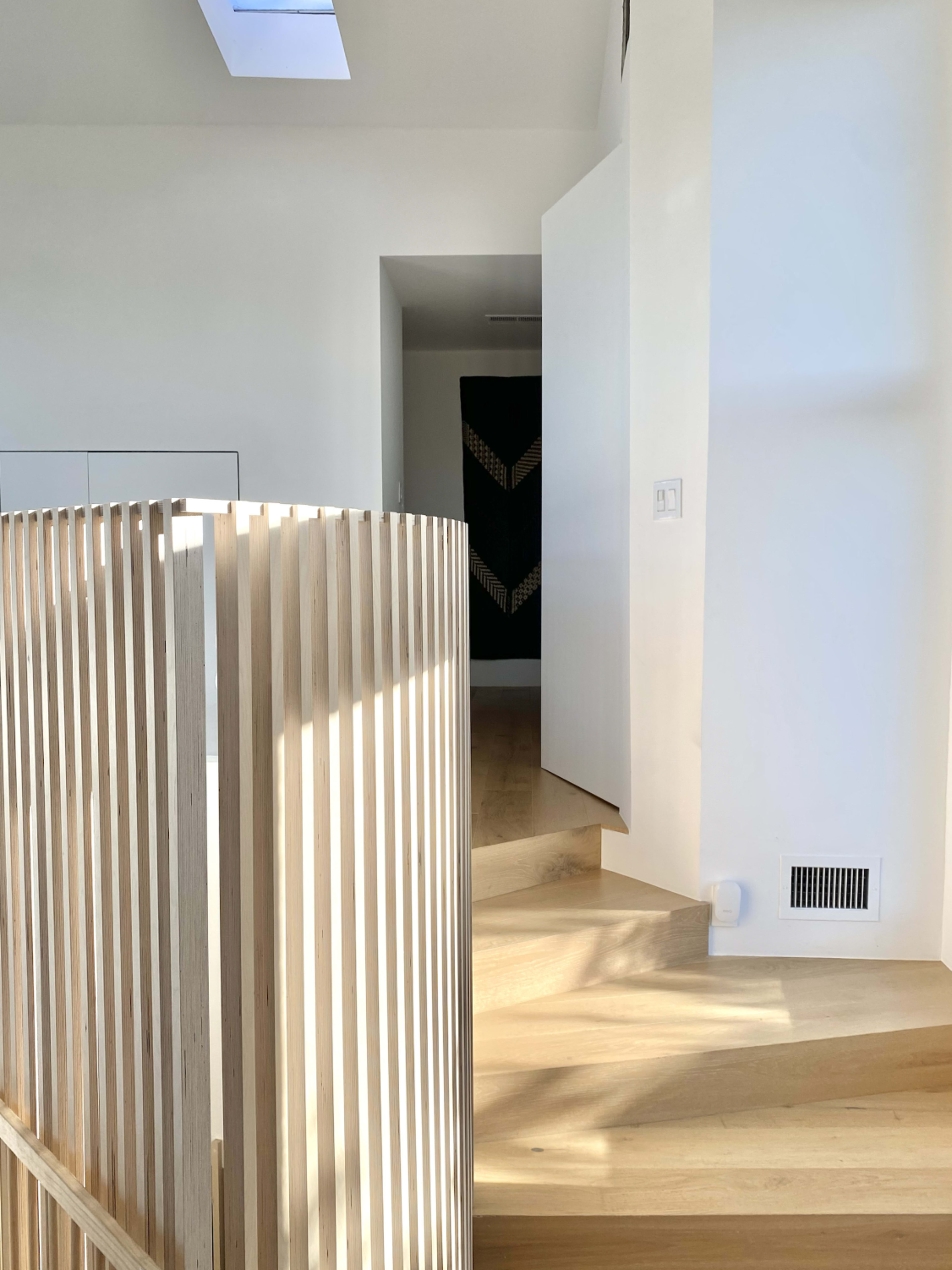 A modern interior space featuring wooden stairs and a slatted wooden railing with a white wall and a doorway in the background.