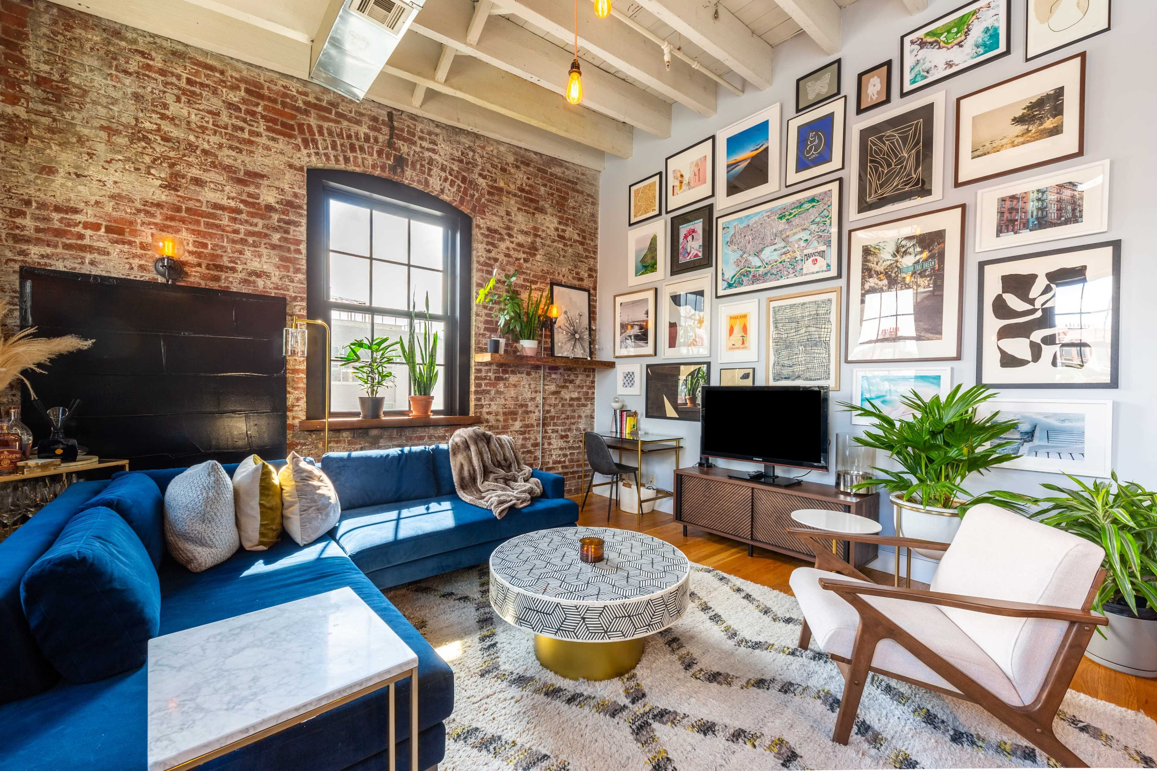 The living room features a blue velvet sofa, a round coffee table, and a gallery wall adorned with various framed artworks against a brick backdrop.