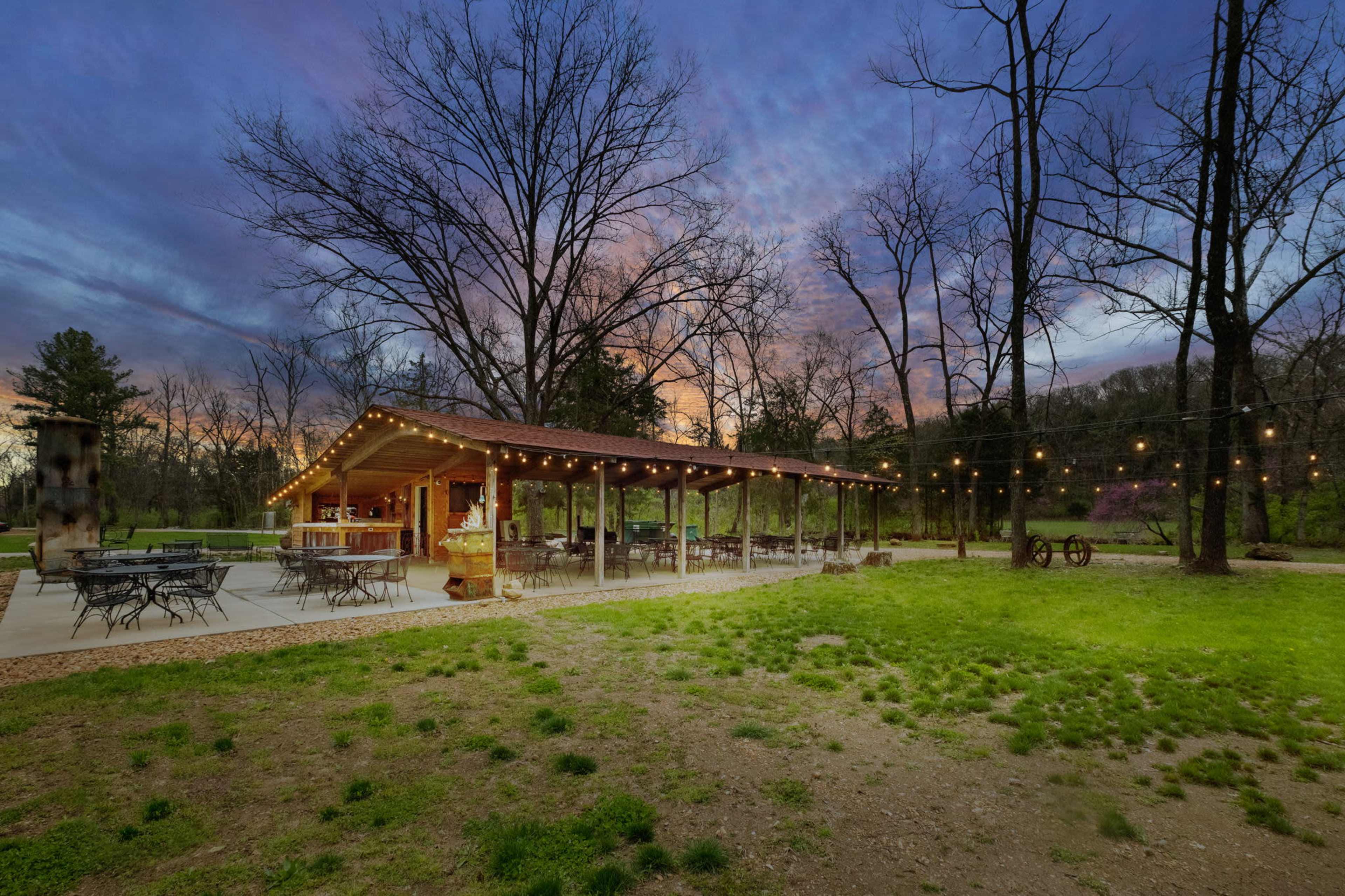 A rustic pavilion with string lights is situated beside a grassy area and surrounded by bare trees during twilight.
