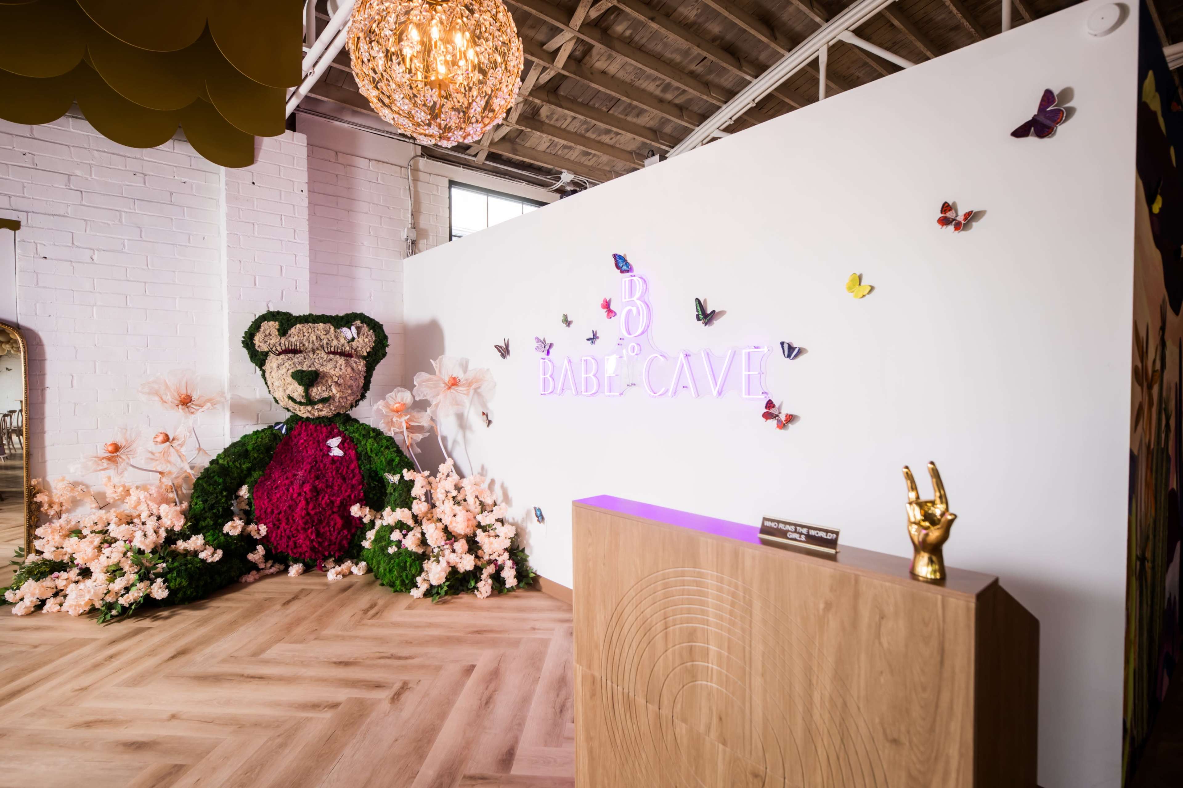 The image depicts a welcoming space featuring a large teddy bear sculpture made of greenery and flowers, alongside brightly colored butterflies and a sign reading "B Babe Cave."