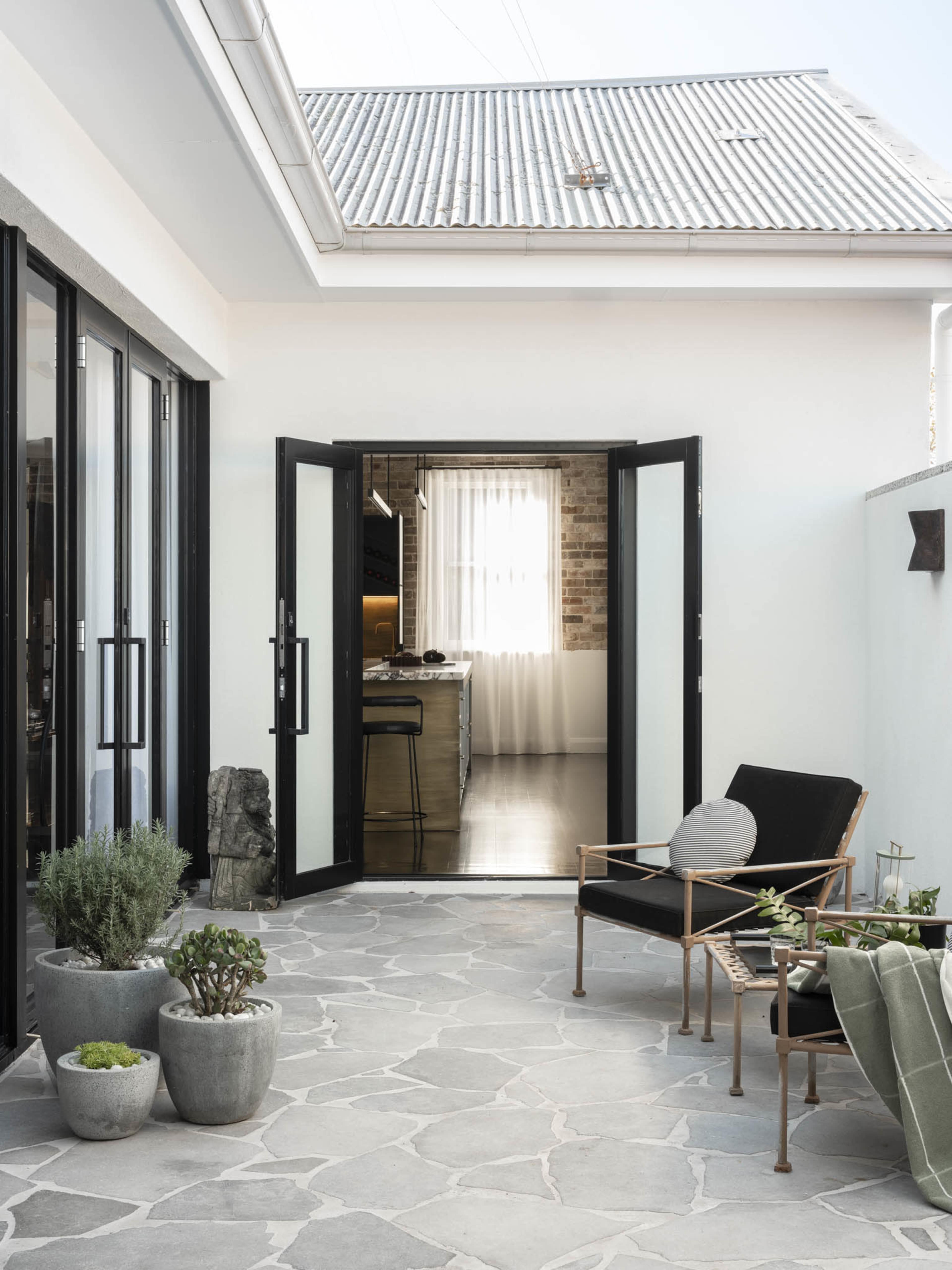 A modern outdoor patio features a stone floor, potted plants, and black-framed doors opening into a bright interior space.