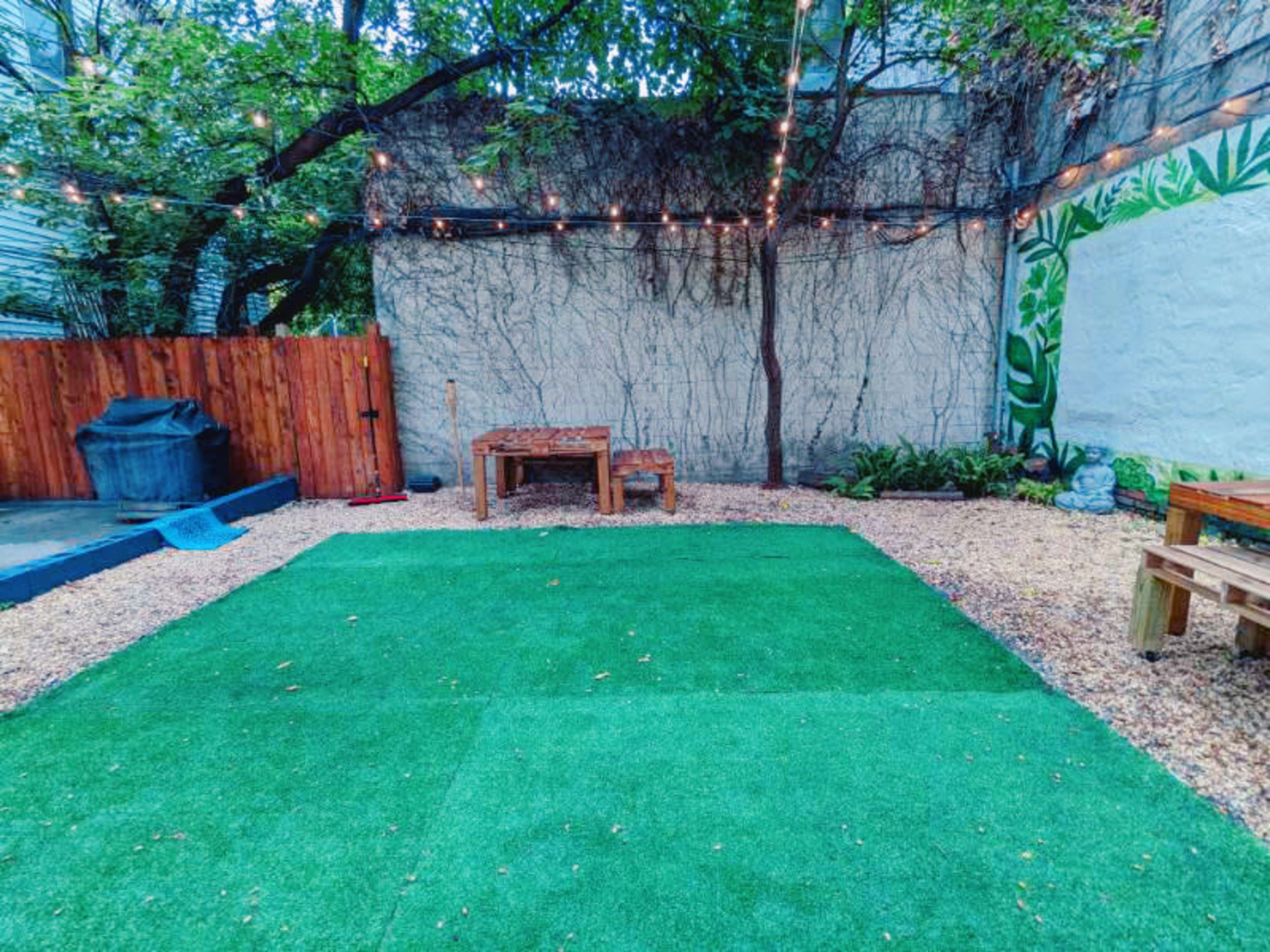 The image shows a landscaped outdoor area with artificial grass, wooden benches, a table, and string lights overhead, surrounded by greenery and a textured wall.