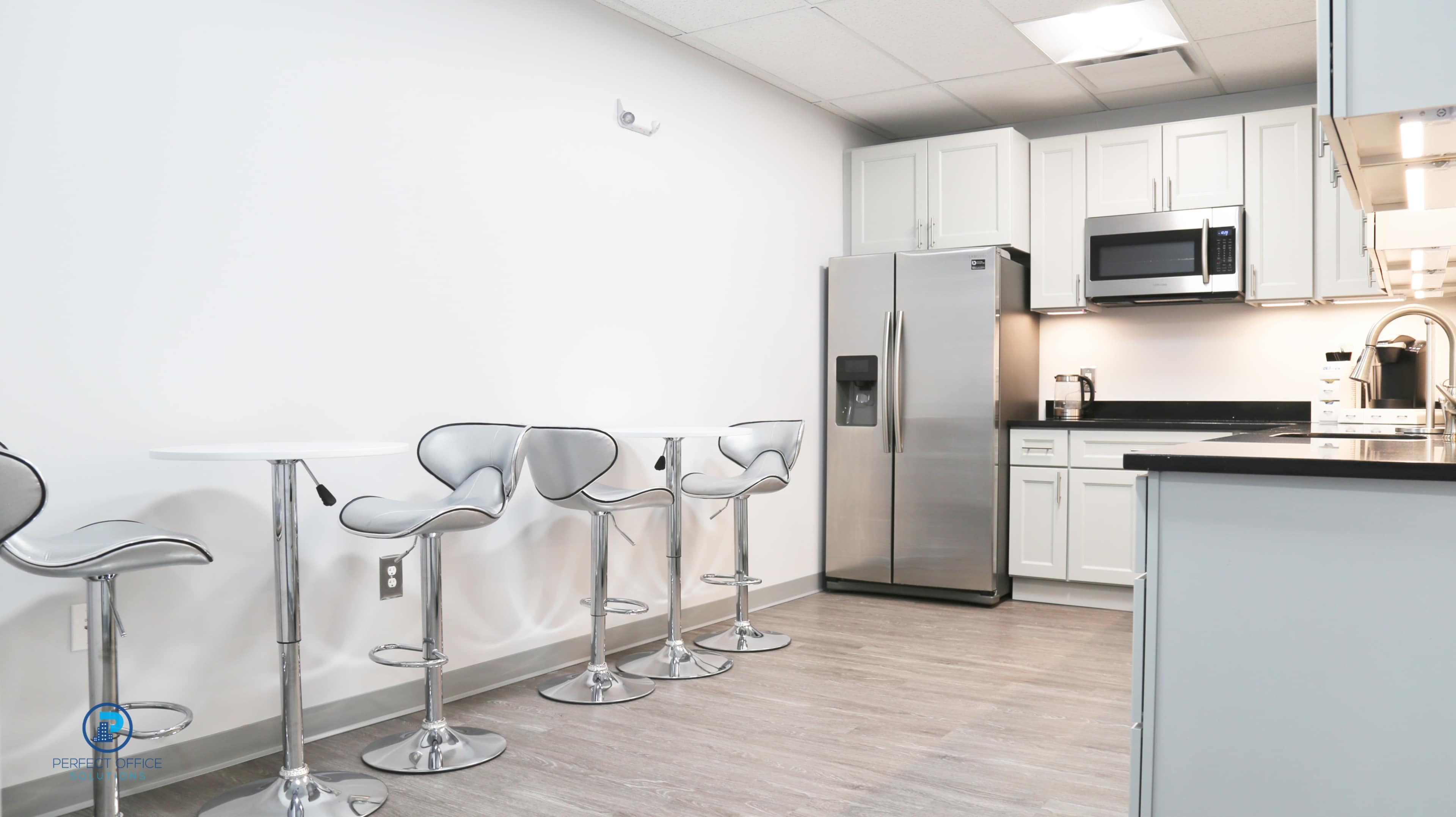 A modern kitchen area with a stainless steel refrigerator, a microwave, and four high-backed chrome stools arranged around two sleek white tables.
