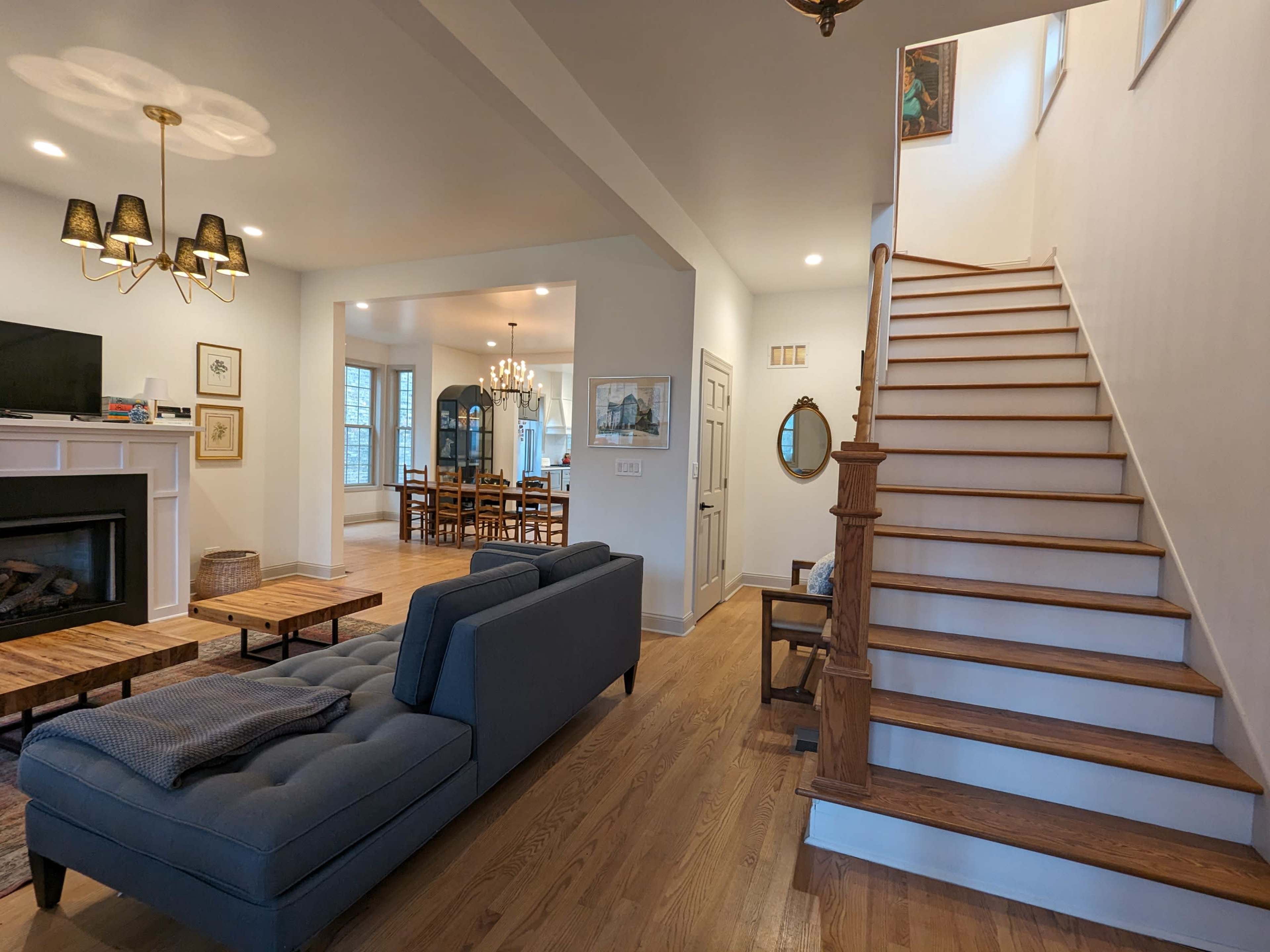 A modern living room leading to a staircase, with a couch facing a fireplace and a dining area visible in the background.