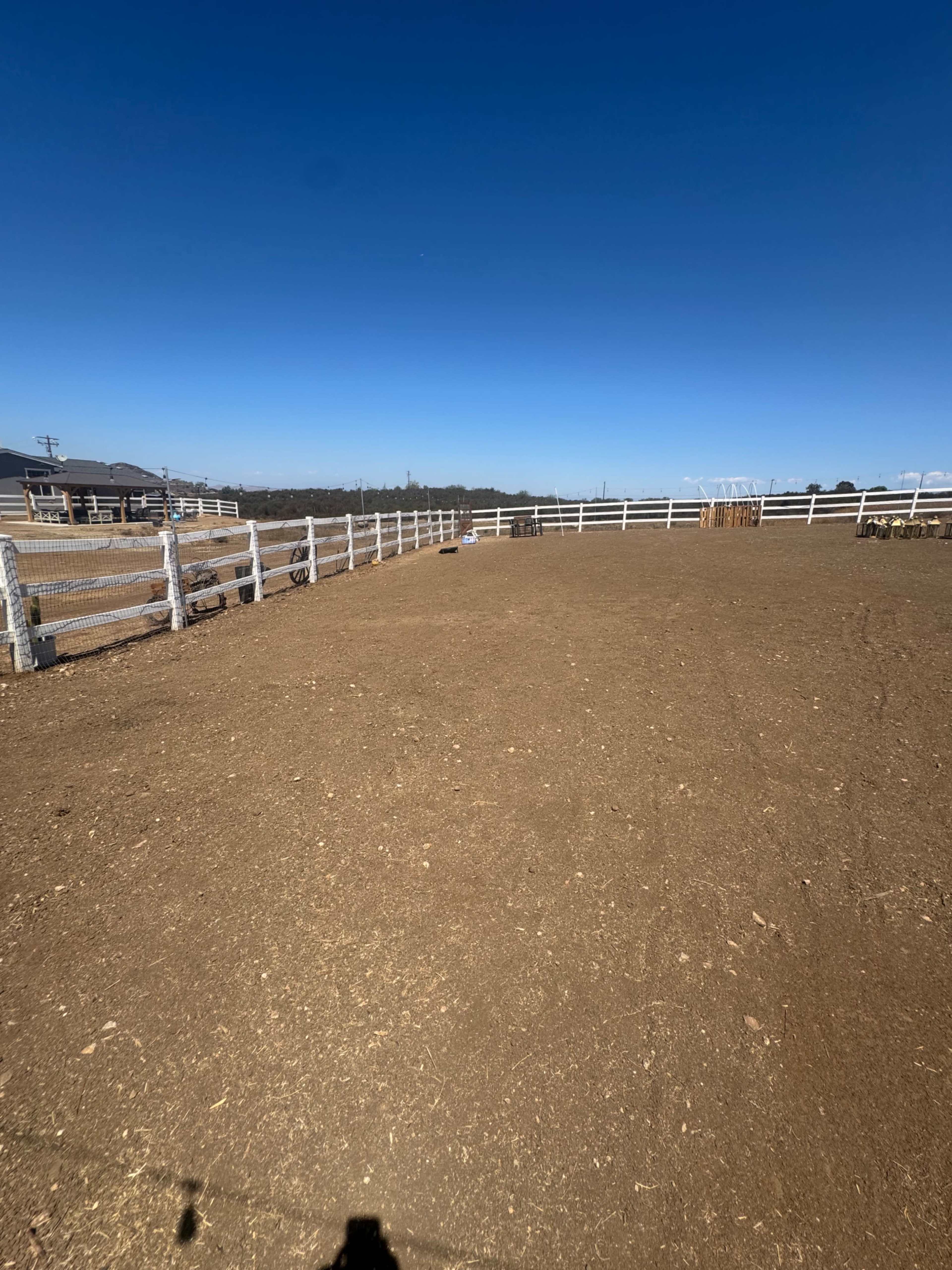 The image shows a large, open area with a dirt surface surrounded by white wooden fencing under a clear blue sky.