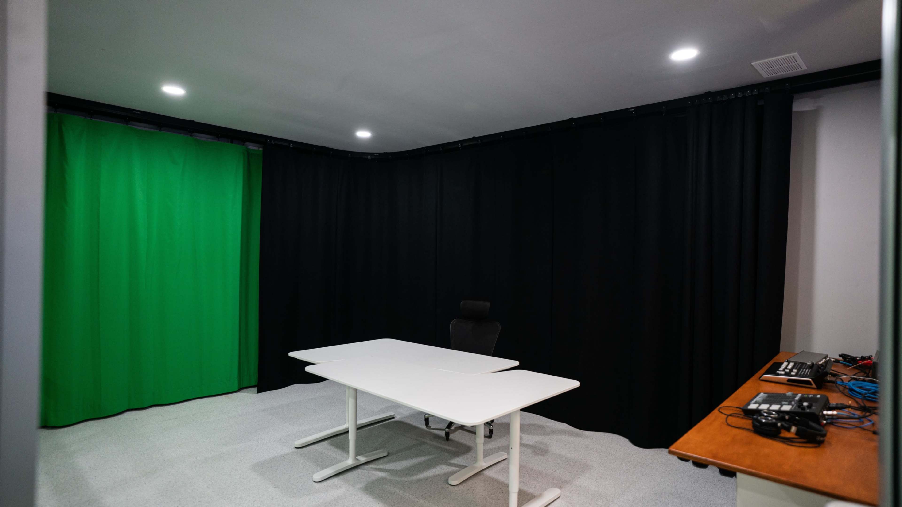 A small studio space with a green screen, black curtains, and a white table with a chair.