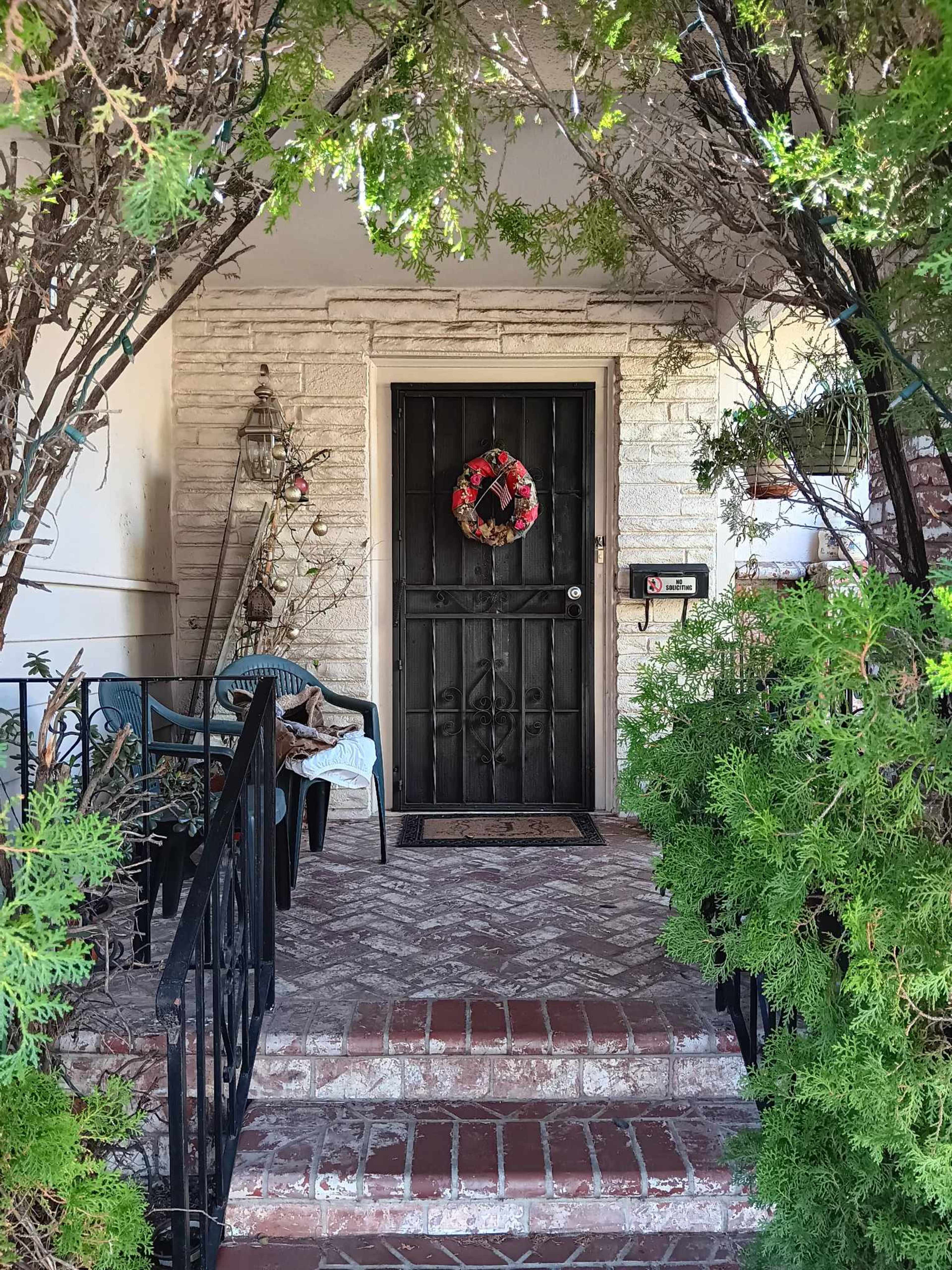 The image shows a porch with a black door adorned with a wreath, surrounded by greenery and two blue chairs.