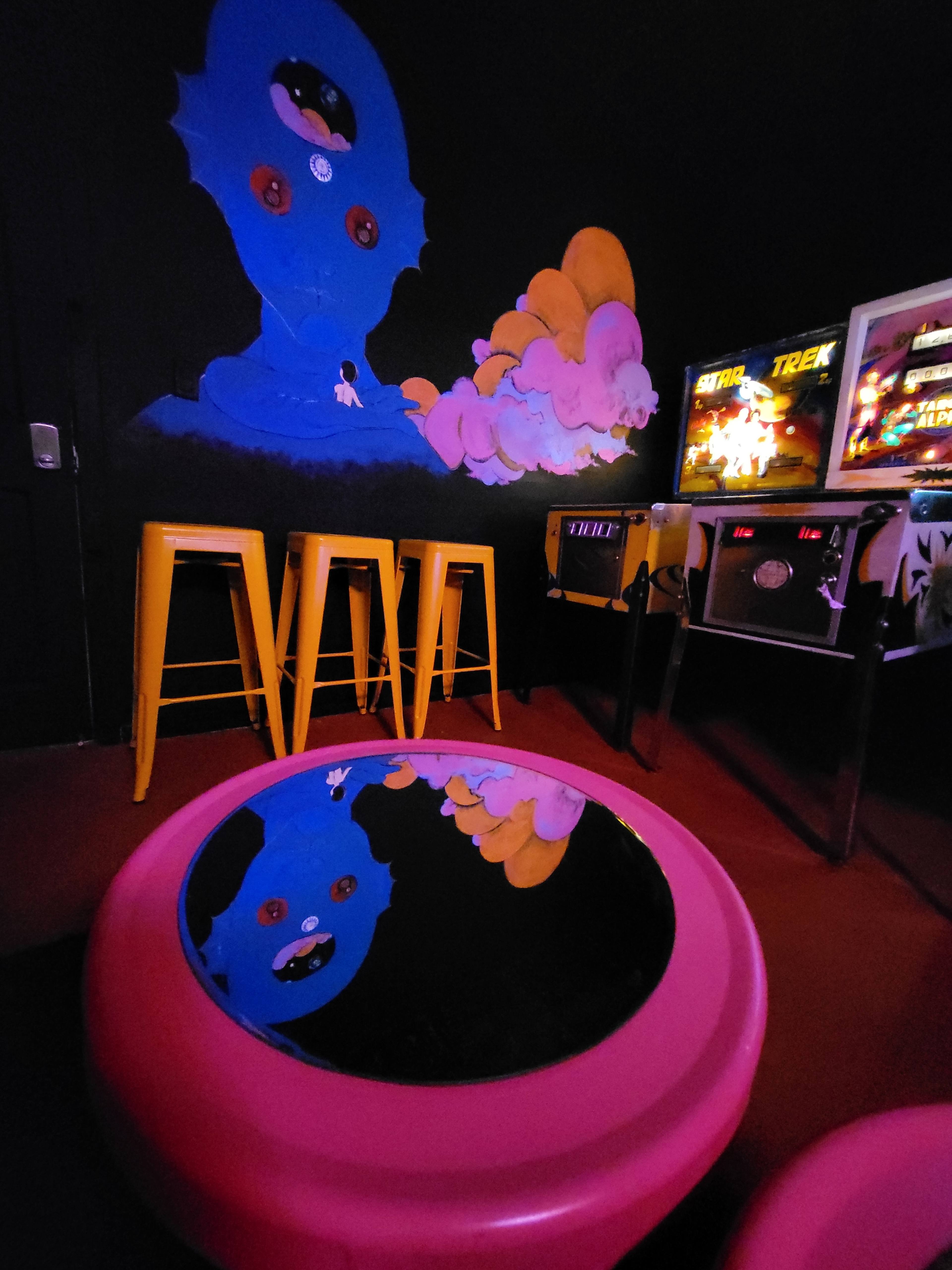 A colorful arcade room featuring a pink circular table with a mirrored surface, yellow stools, and vintage arcade machines against a black wall adorned with blue and orange artwork.