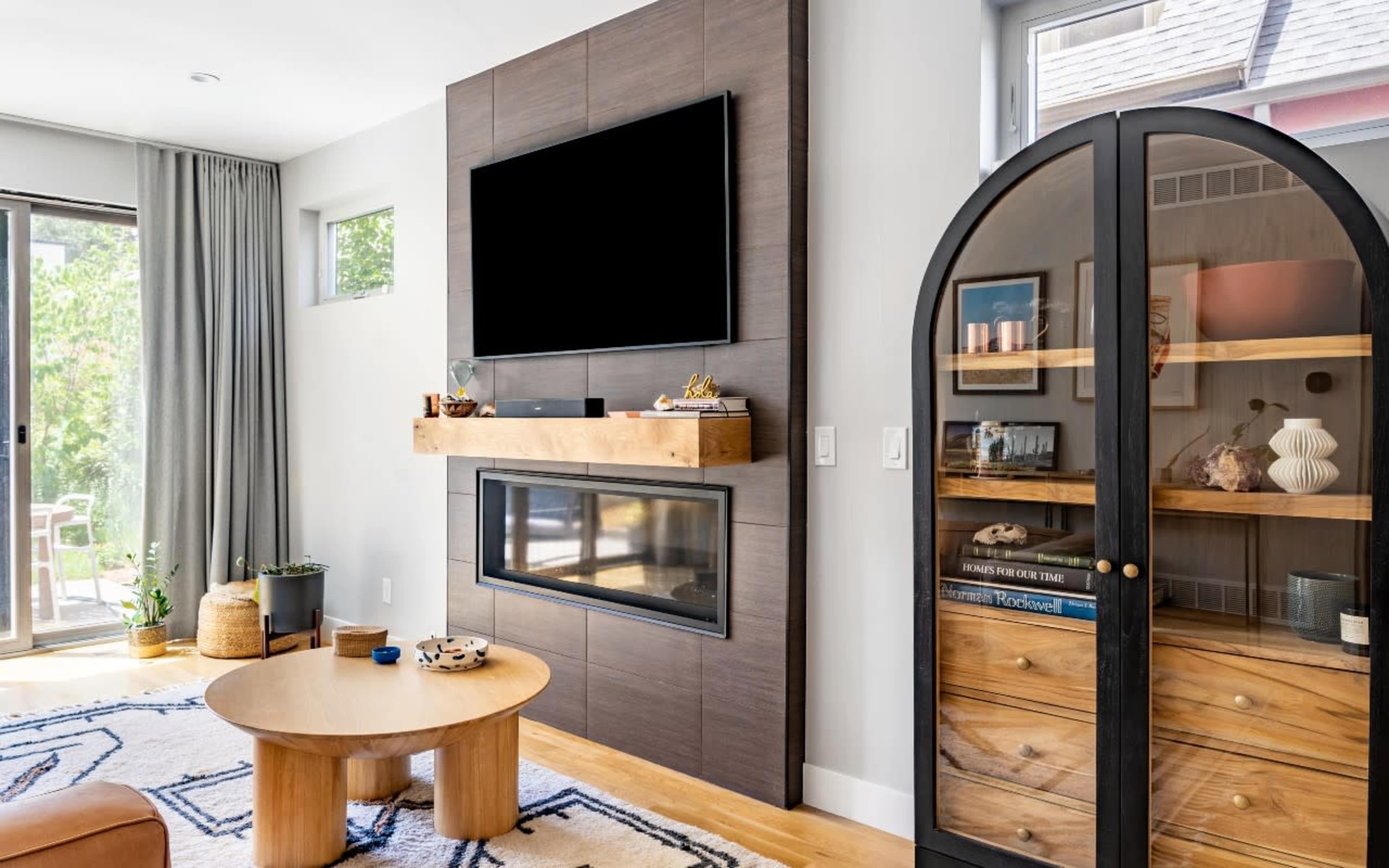 The image shows a modern living room with a wall-mounted television above a wooden fireplace and a curved glass-front cabinet displaying decorative items.