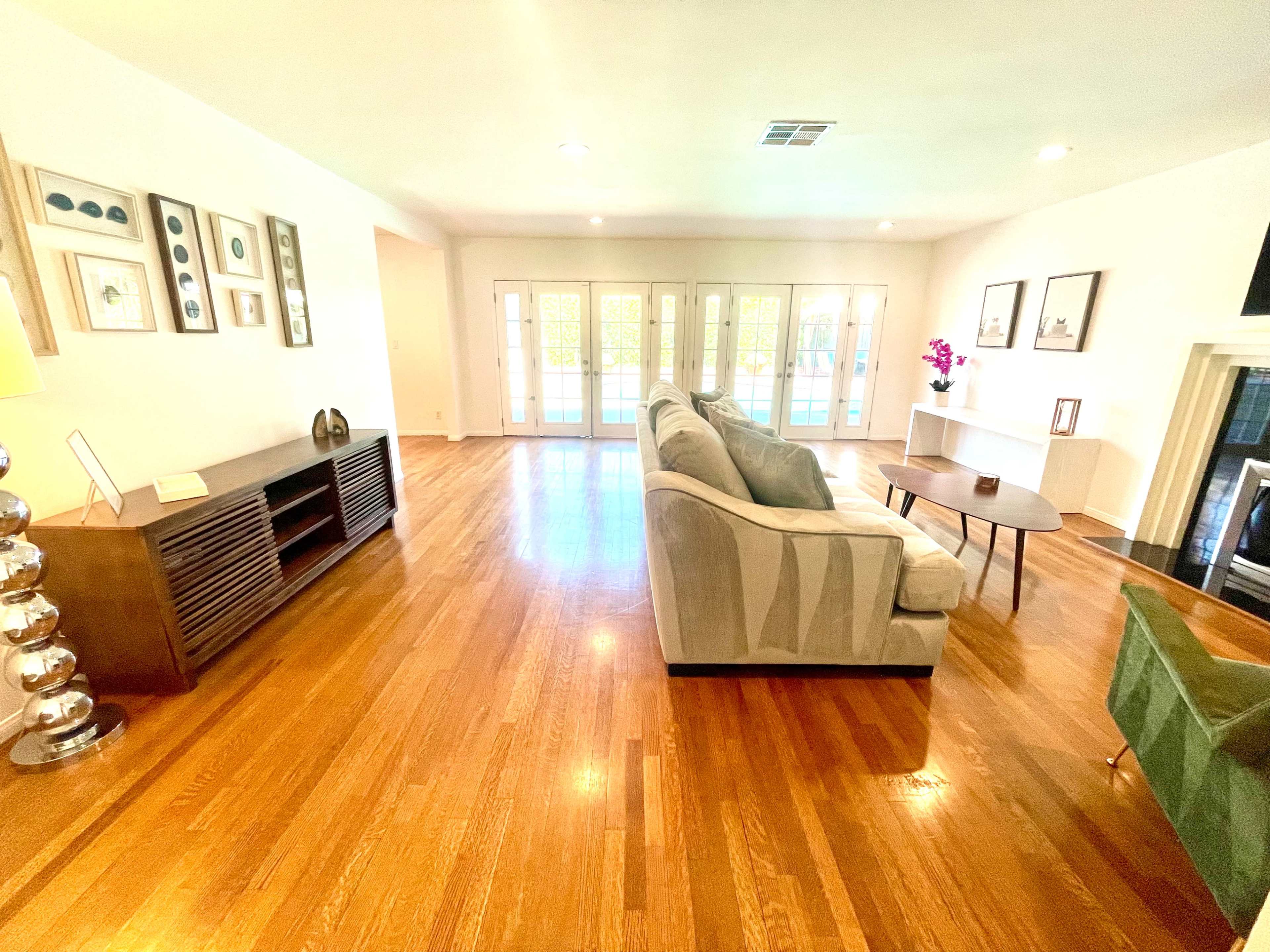 A spacious living room with polished wooden floors, a large sofa, a coffee table, and a console table, all illuminated by natural light coming through multiple glass doors.