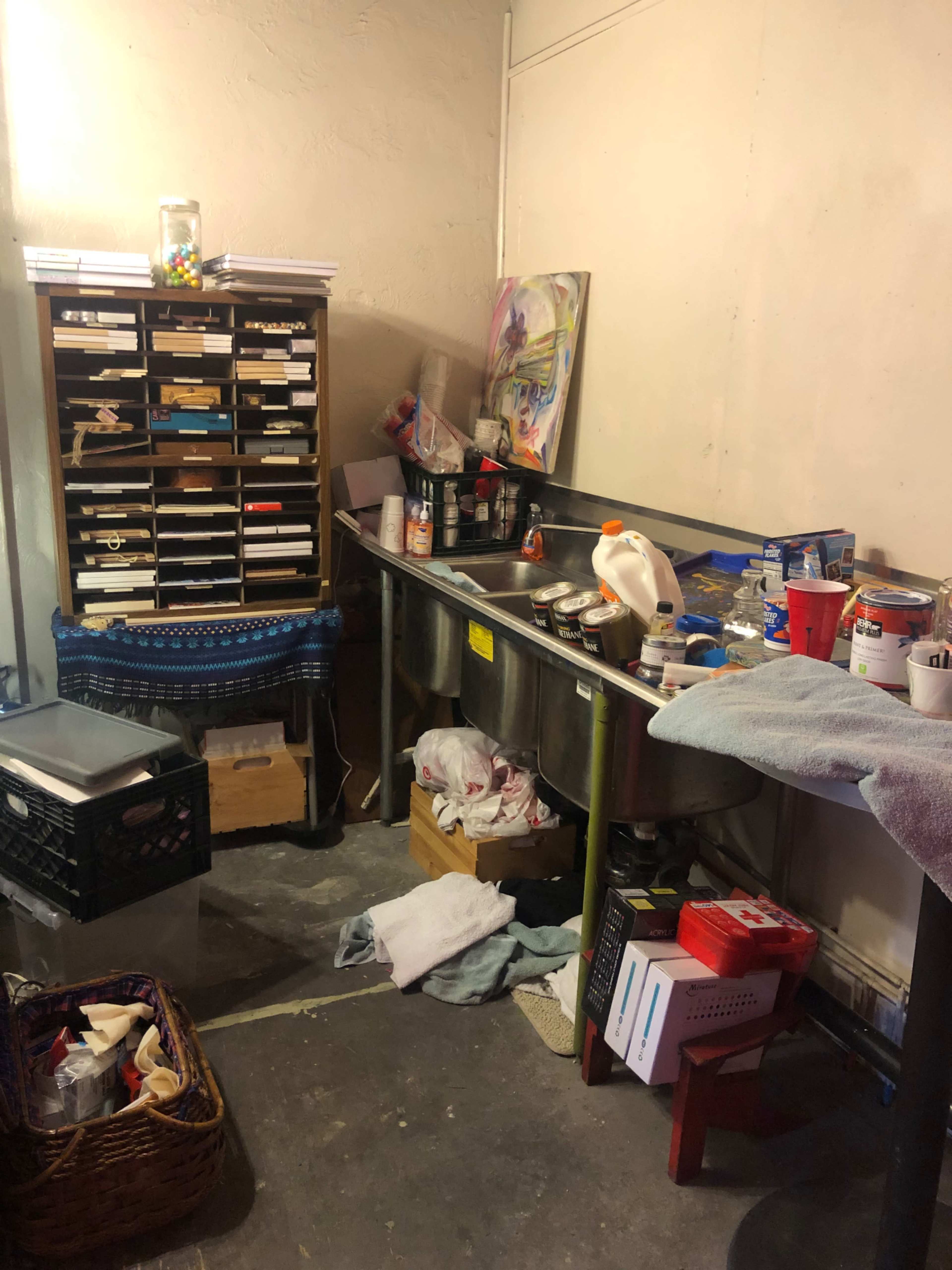 A cluttered workspace with a stainless steel sink, various containers, and a storage unit filled with boxes and supplies.