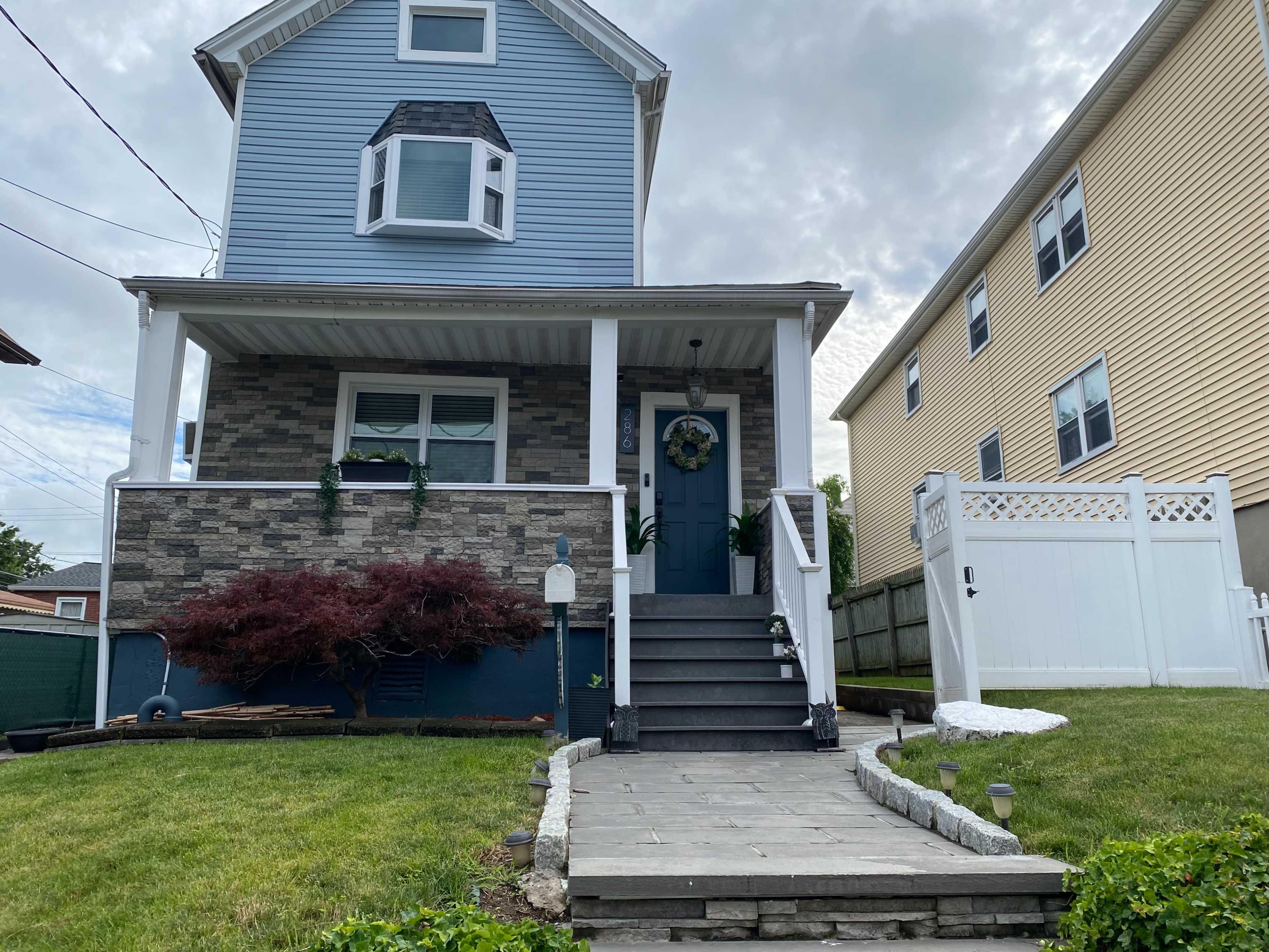 A two-story blue house with stone accents features a front porch, a set of stairs, and a well-maintained lawn, bordered by a white fence.