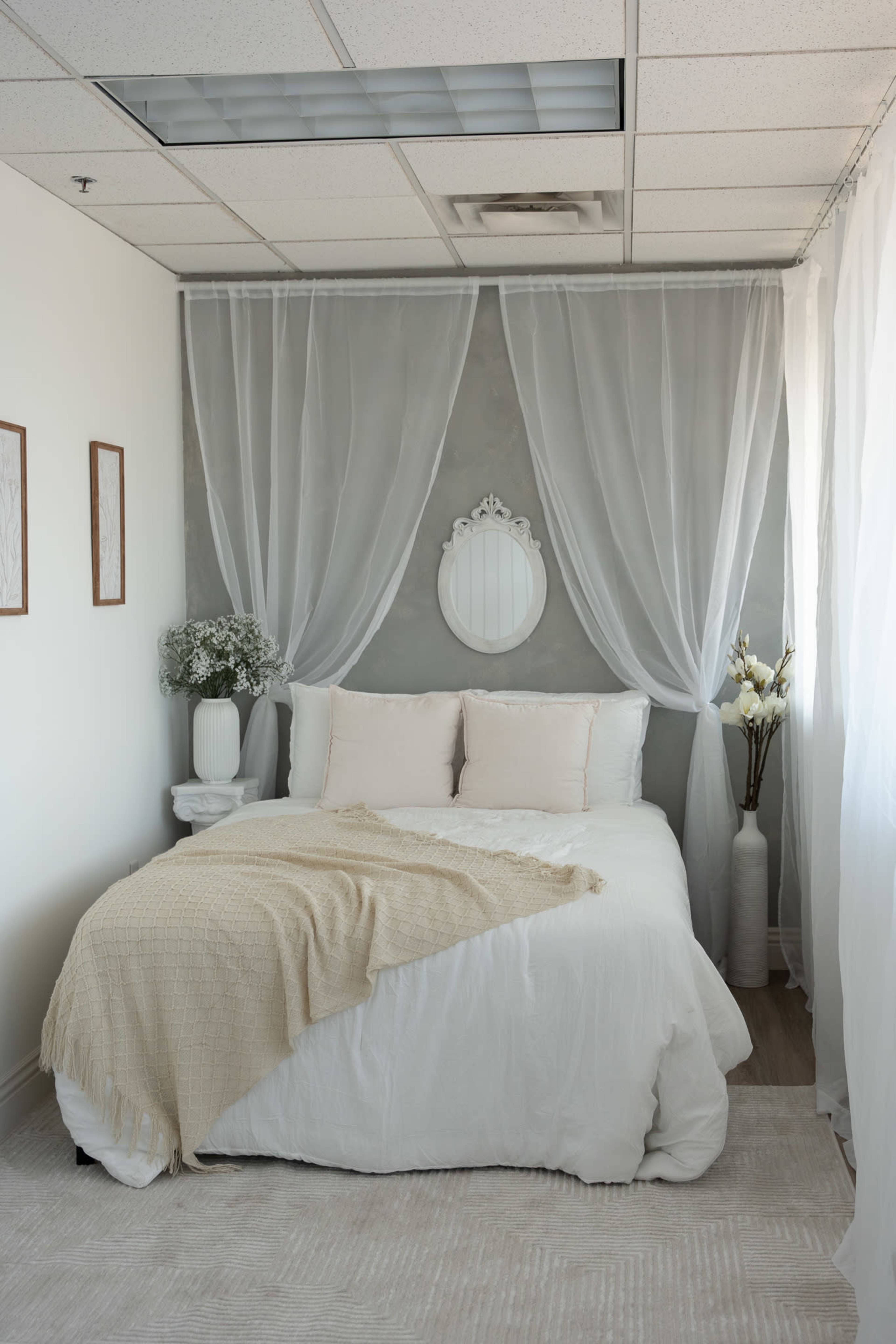 The image shows a neatly arranged bedroom with a white bed adorned with light-colored pillows and a cozy blanket, framed by sheer curtains and minimalistic decor.