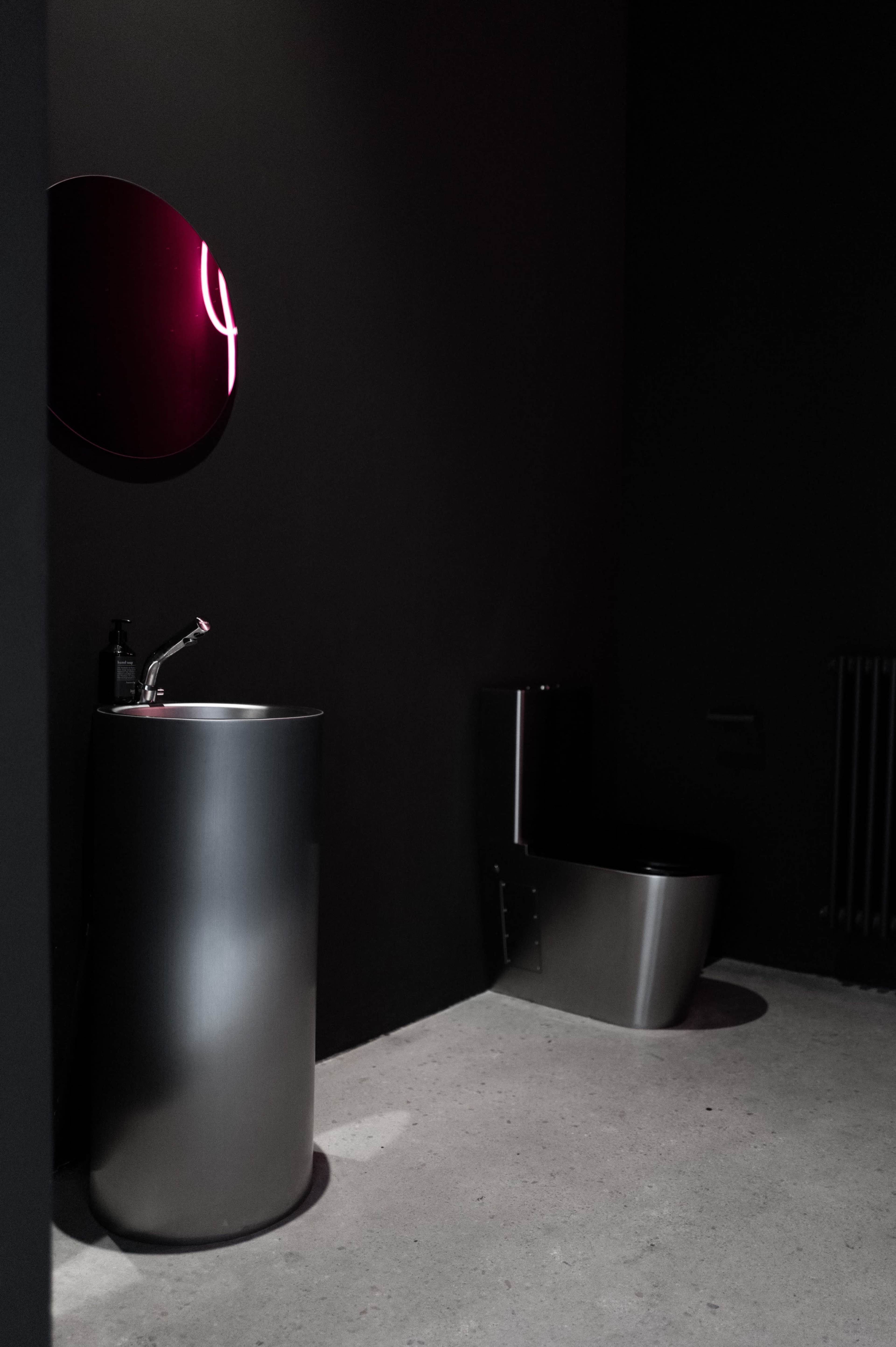 The image shows a modern bathroom with a sleek round sink and a minimalist toilet against a dark wall.